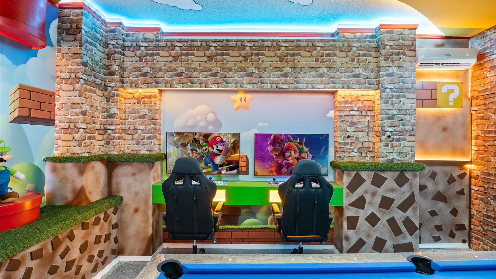 A vibrant Super Mario Bros.-themed game room features brick-patterned walls and a light blue ceiling with cloud murals. Two gaming stations with monitors and black gaming chairs are built into a green platform, surrounded by themed props.