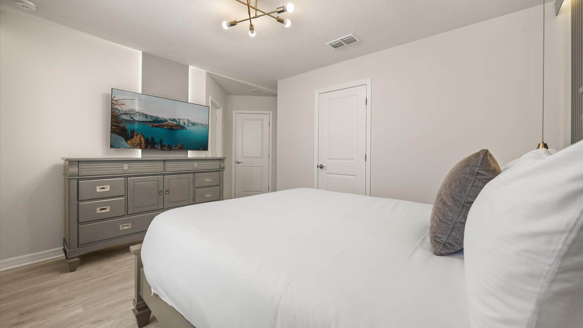 Another angle of the sophisticated bedroom, displaying a grey wooden dresser with a wall-mounted TV, and a view towards a white door and closet.