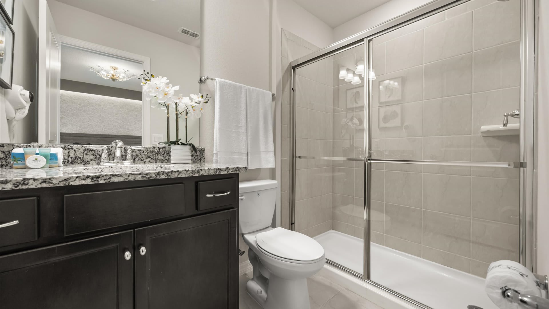 A clean bathroom with a dark wood vanity, speckled granite countertop, white toilet, and a glass-enclosed shower with light beige tiled walls, accented by white orchids.