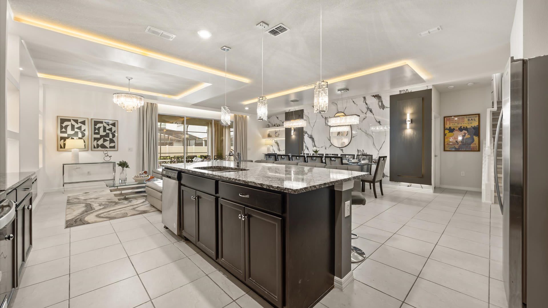 Wide view of the open-concept kitchen and living area, showcasing a large kitchen island with granite countertops and bar seating, dark cabinetry, and the adjacent living space with accent lighting.