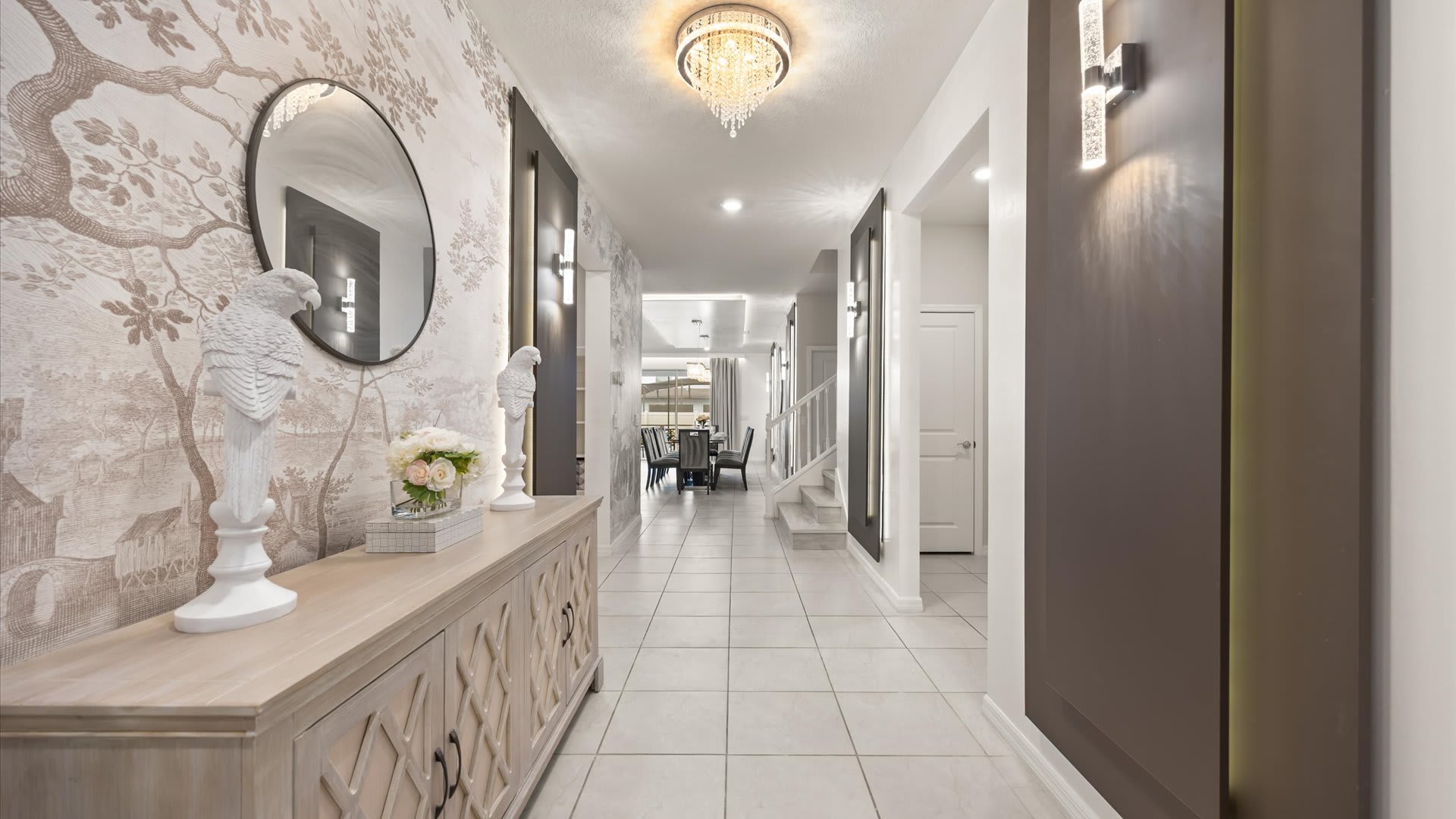 Inviting foyer with light-colored tile flooring, a console table adorned with decorative white bird statues, a round mirror, and a patterned wallpaper accent wall, leading into the main living areas.