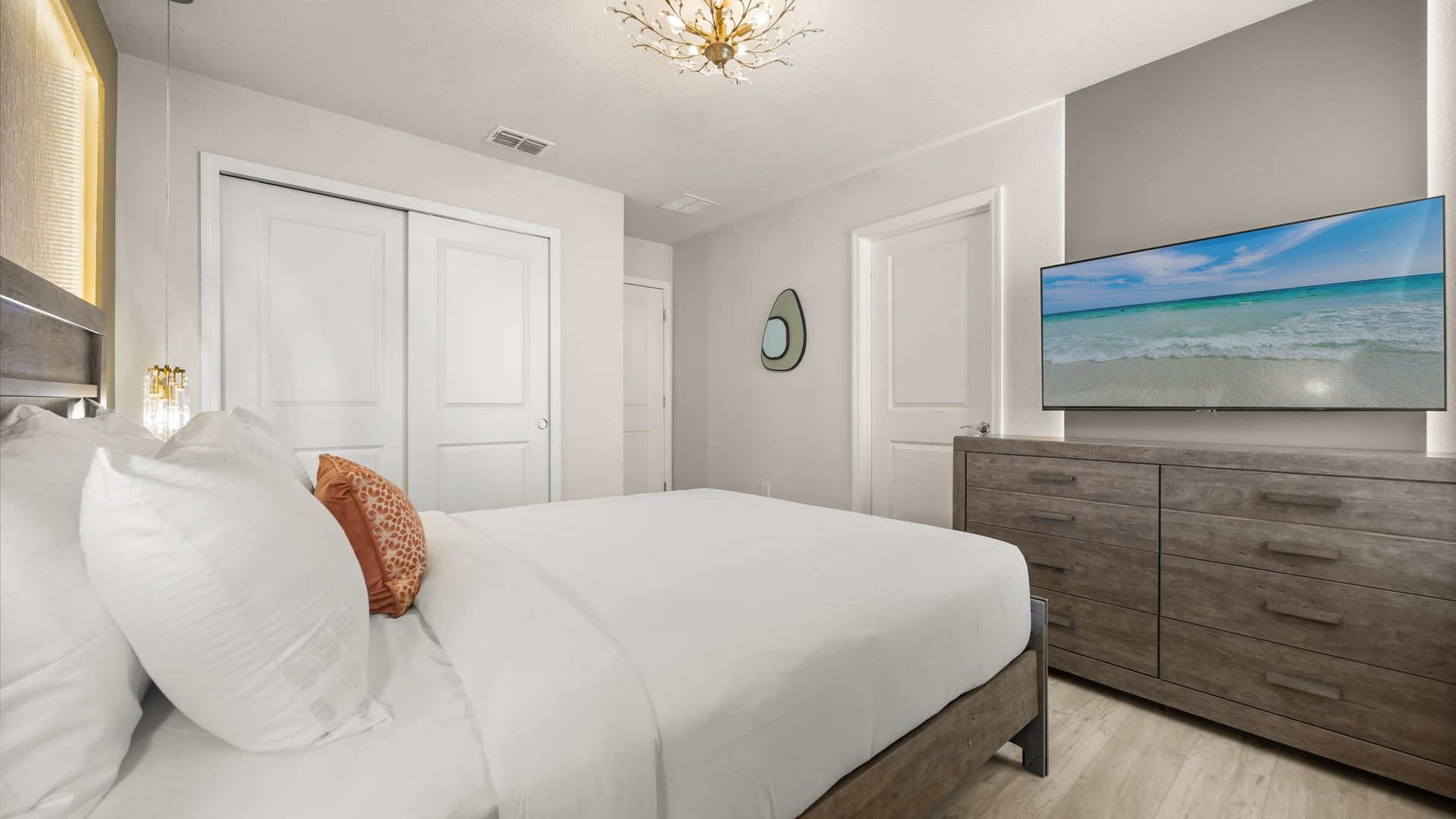 Another view of the stylish bedroom, showing a large grey wooden dresser with a wall-mounted TV displaying a beach scene, and a closet with white sliding doors.
