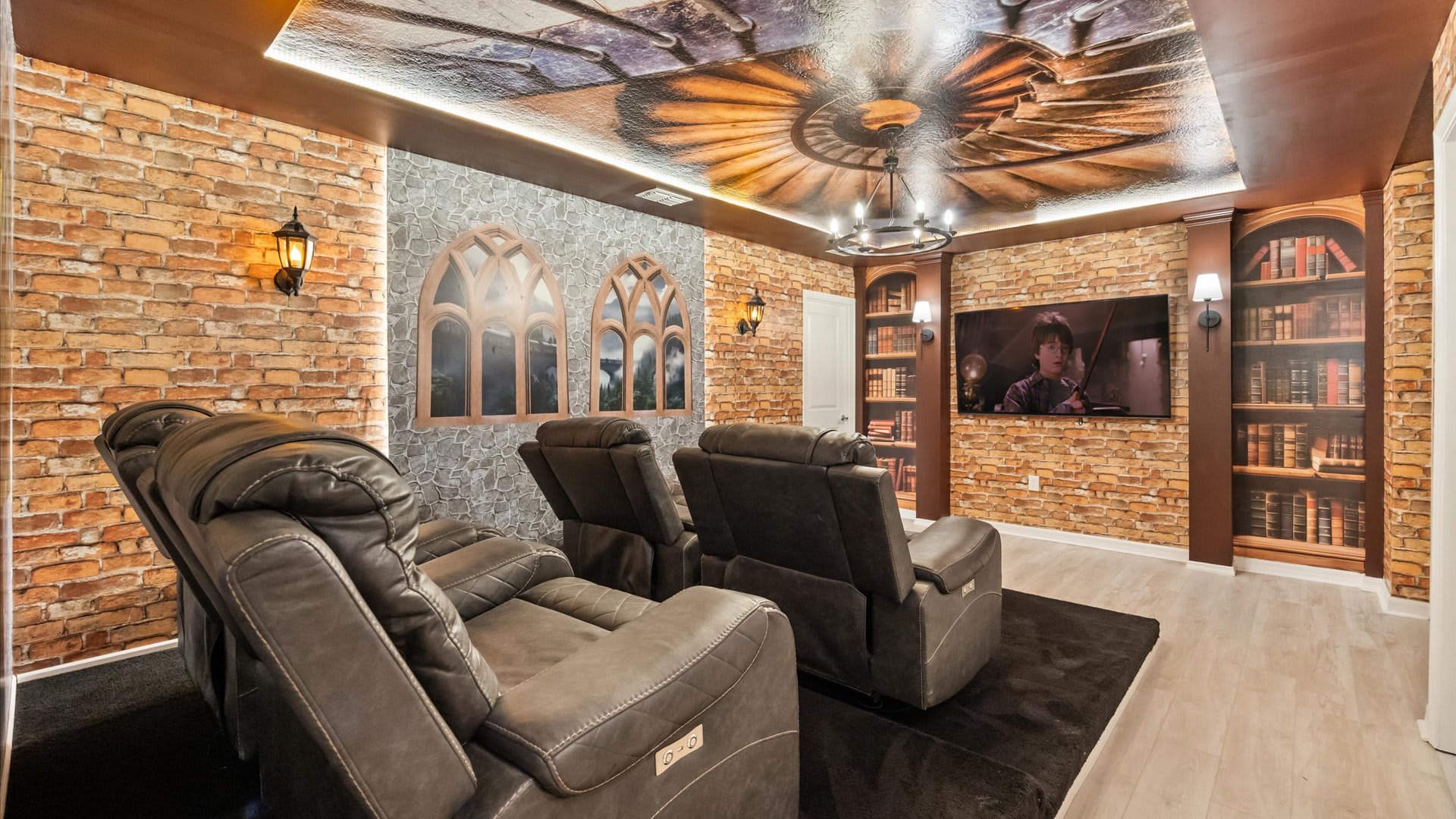 A Harry Potter-themed media room features a large TV flanked by faux bookshelves, brick-patterned walls, and a ceiling mural depicting a grand staircase. Four plush, dark gray reclining chairs face the screen, set on a dark rug.
