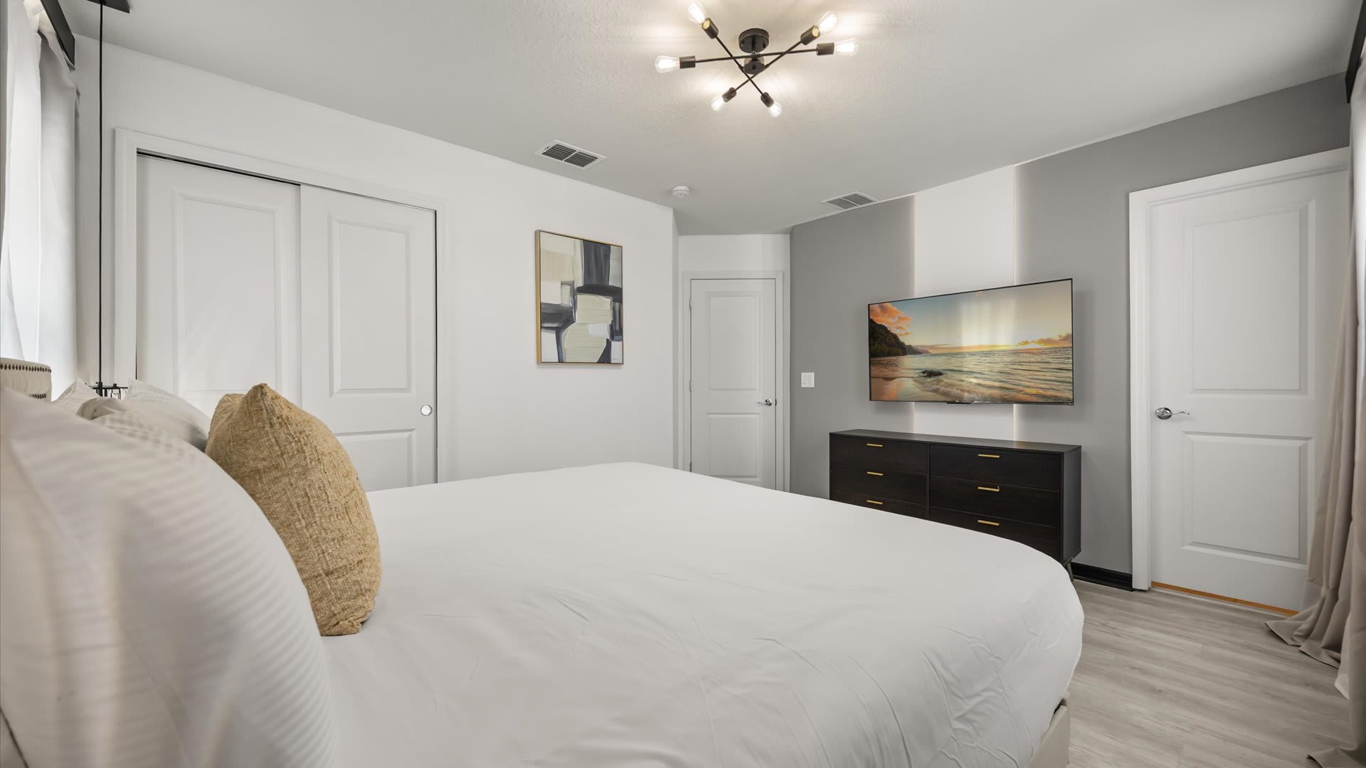 Bright bedroom with a large white bed, a dark wood dresser, and a wall-mounted TV displaying a beach scene, featuring a gray accent wall behind the TV.