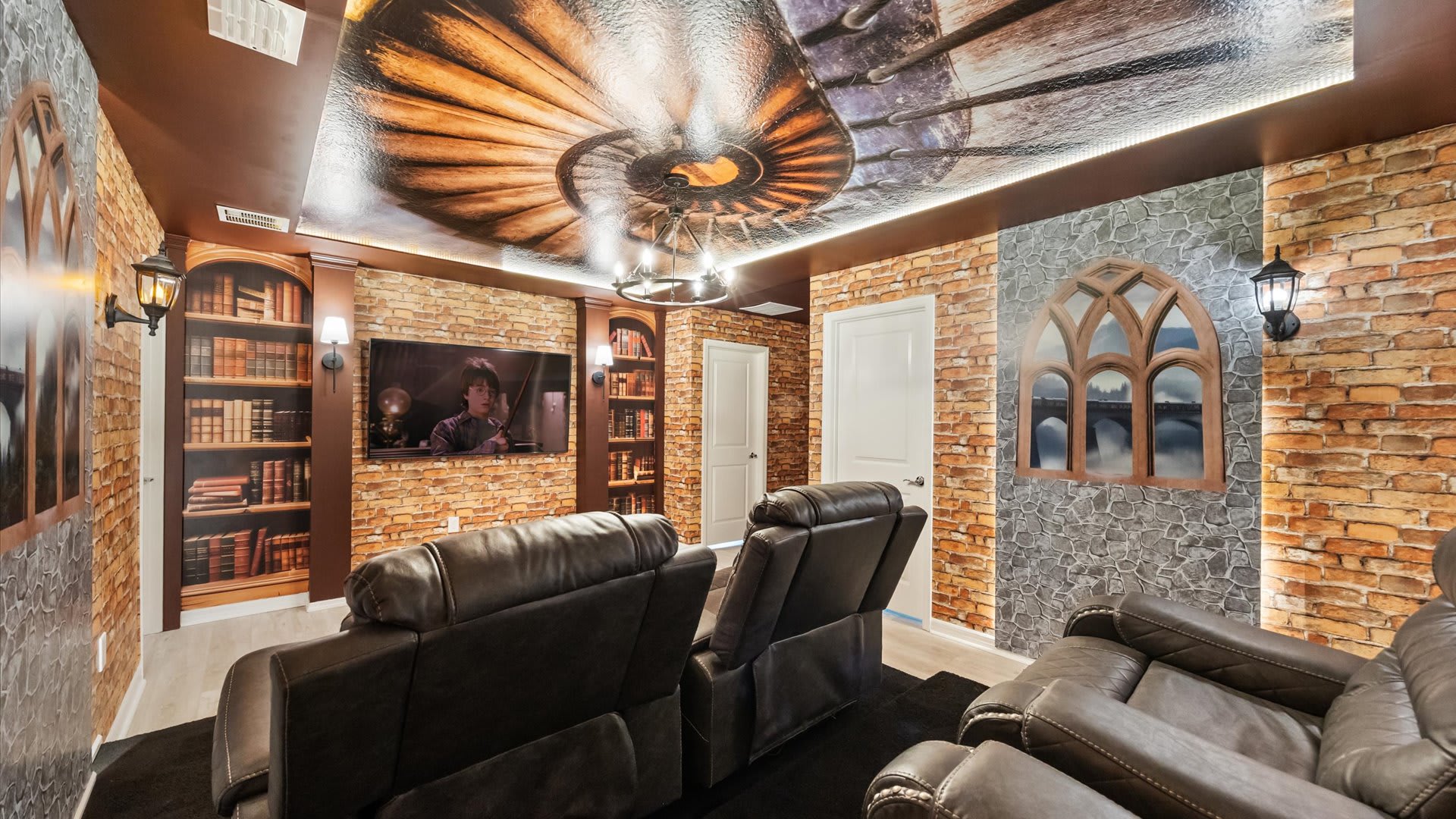 Themed media room with comfortable leather recliners facing a large wall-mounted TV, set against a brick and stone wall backdrop. The ceiling features a spiral design, and built-in bookshelves flank the TV, creating a magical ambiance.