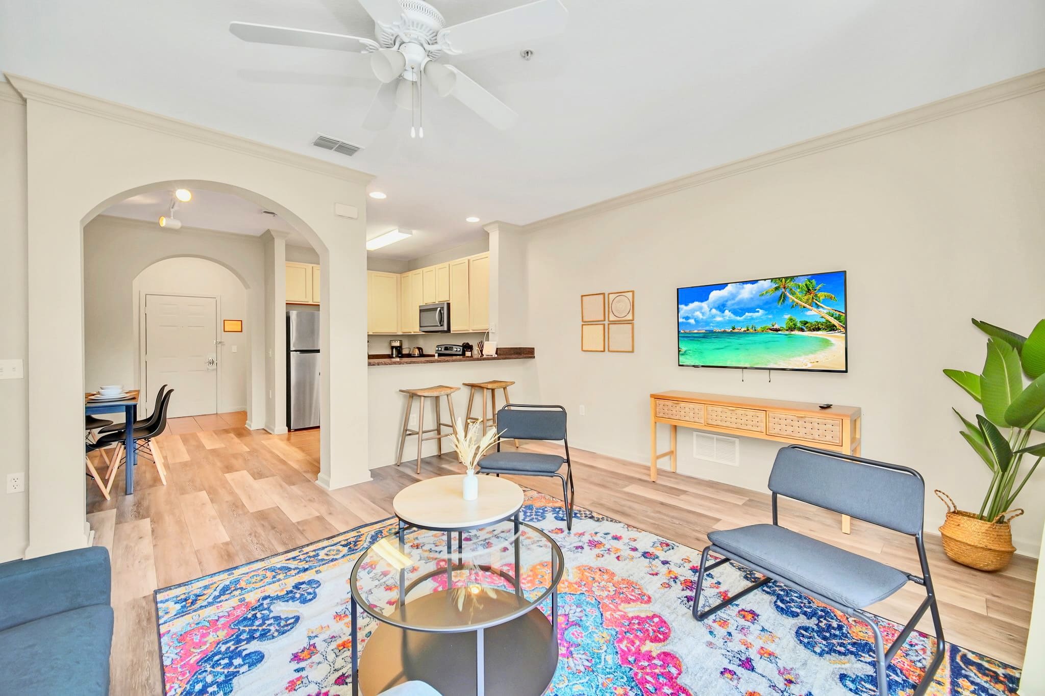 Open-concept living room with a colorful area rug and wood-look flooring, seamlessly transitioning into a kitchen area with a breakfast bar and light-colored cabinets. A wall-mounted TV is visible, enhancing the modern aesthetic.