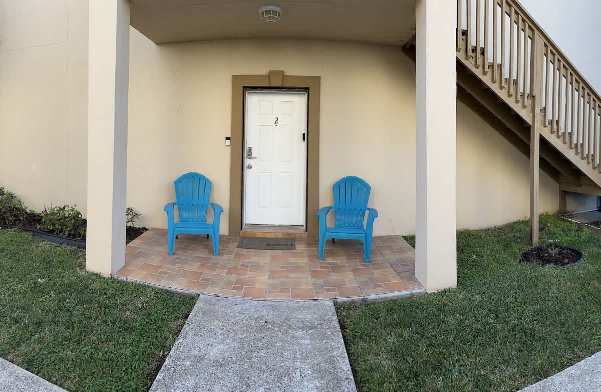 The front entrance of the condo, featuring a white door with the number "2" and two bright blue Adirondack chairs on a tiled patio. A concrete walkway leads to the entrance, surrounded by green grass.