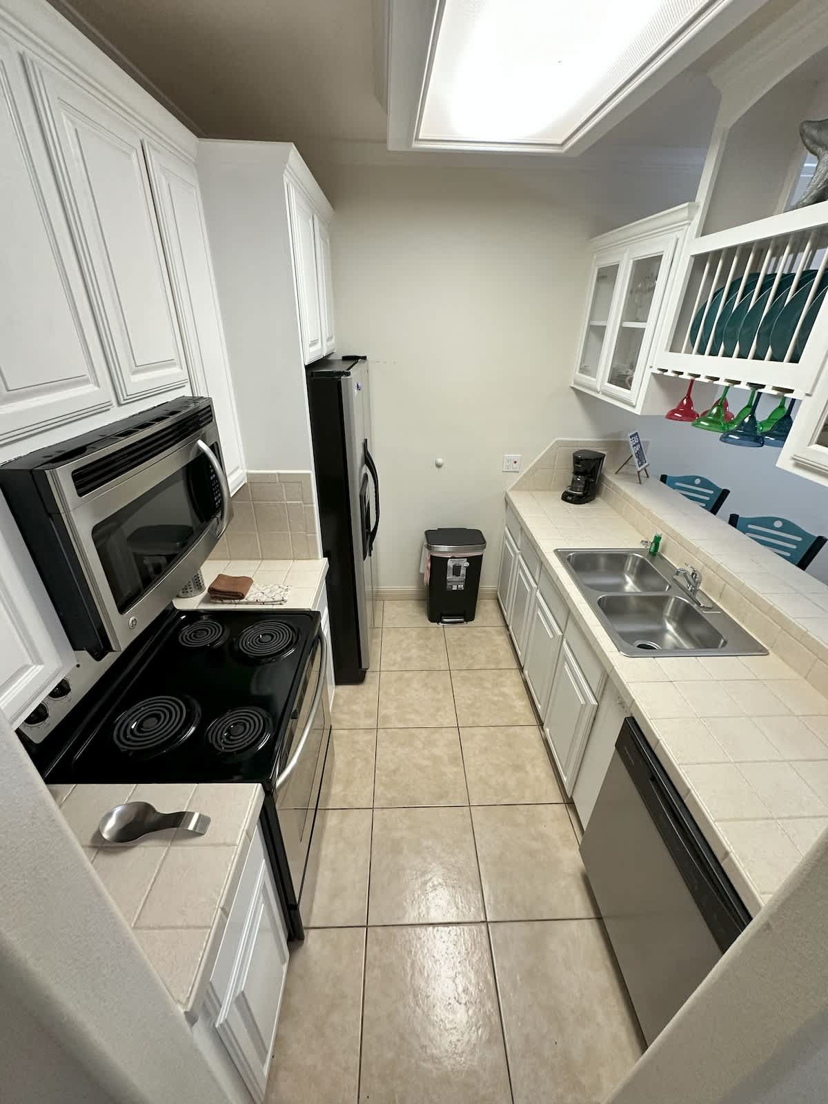 A galley kitchen with white upper and lower cabinets, light tile countertops, and stainless steel appliances including a microwave, stove, and refrigerator. A double sink is visible on the right, and a black trash can is placed in the middle.