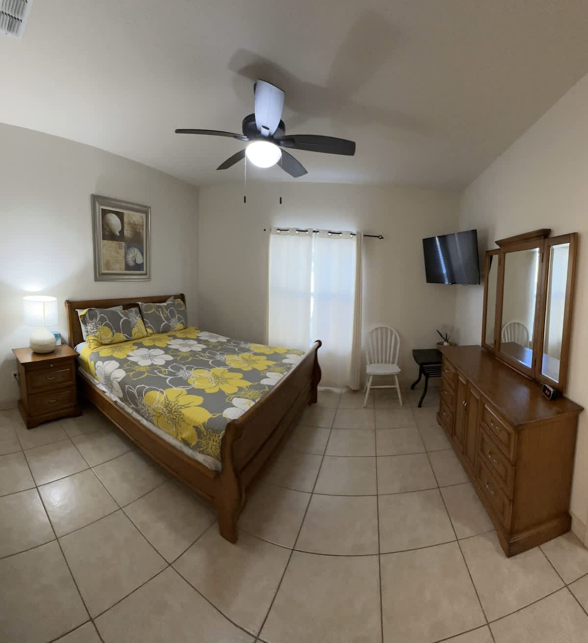 A bright bedroom featuring a wooden sleigh bed with yellow and gray floral bedding, a matching wooden dresser with a triple mirror, and a wall-mounted TV. A ceiling fan with lights hangs overhead, and a white chair is visible near the window.