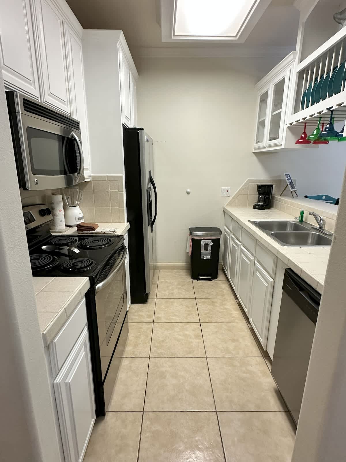 A narrow galley kitchen featuring white cabinetry, light tile countertops, and stainless steel appliances including a microwave, stove, and refrigerator. A coffee maker is visible next to the double sink, and a dishwasher is integrated below.