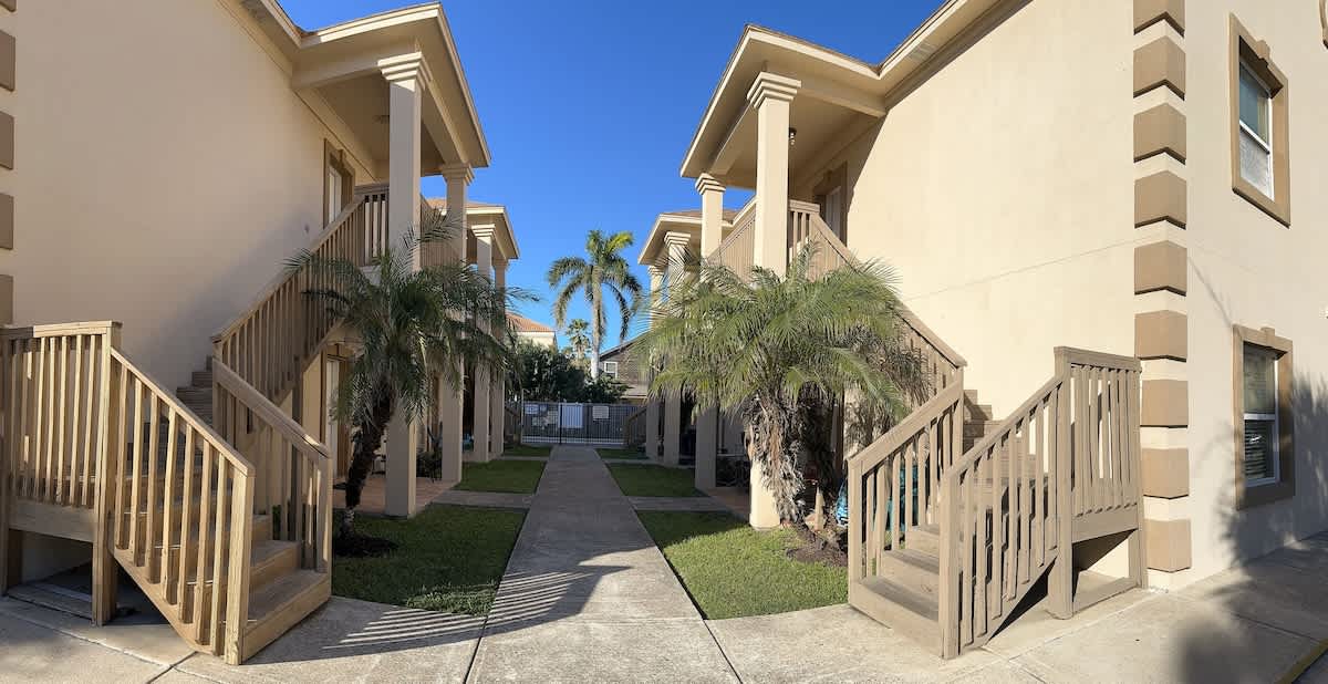 An exterior view of a multi-unit property, showing two-story buildings with light-colored stucco facades and wooden staircases leading to upper units. A paved walkway is flanked by green grass and palm trees under a clear blue sky.