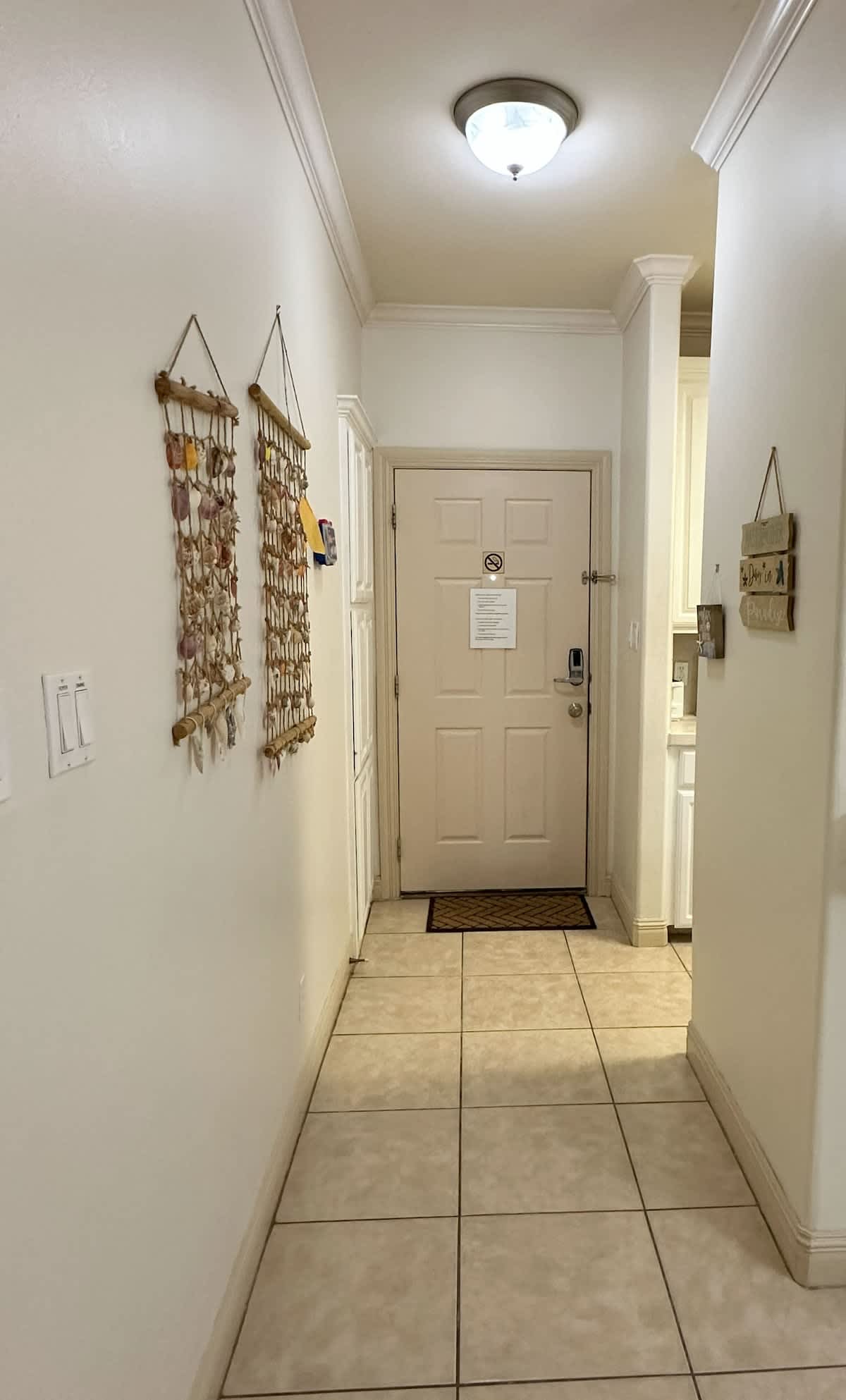 A narrow foyer with light tile flooring and white walls, leading to the main entrance door. Decorative shell and driftwood hangings adorn the left wall, and a small mat is placed in front of the door.
