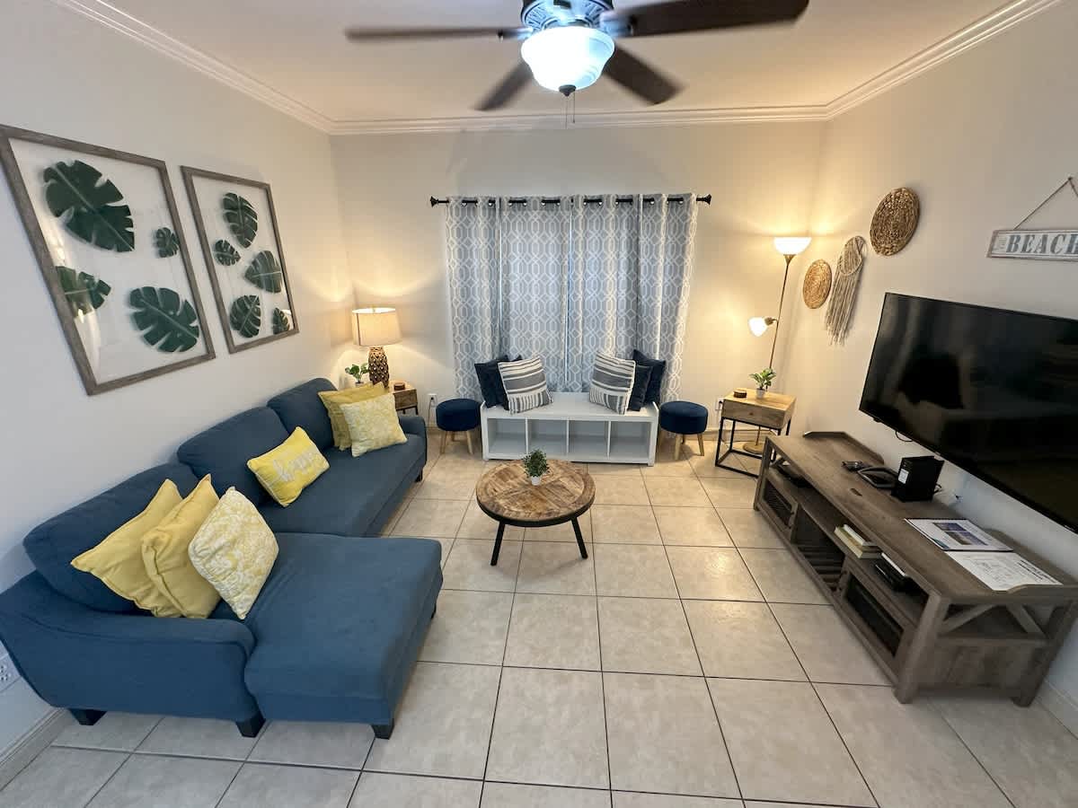 A wide-angle view of the living room with light tile flooring, a blue sectional sofa with yellow throw pillows, and a round wooden coffee table. A large flat-screen TV sits on a rustic media console, and patterned curtains cover a window.
