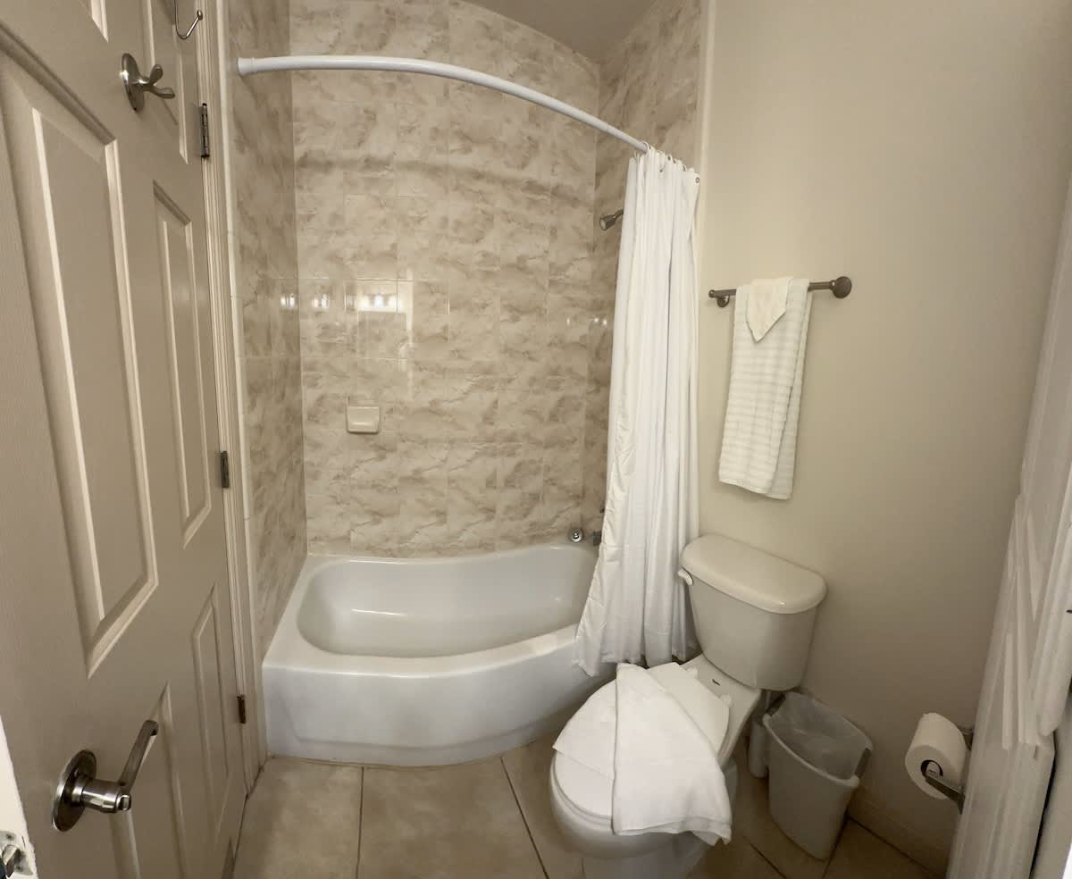 A bathroom featuring a white bathtub with a shower curtain, a toilet, and light-colored tiled walls in the shower area. A towel rack with white towels is mounted on the wall.