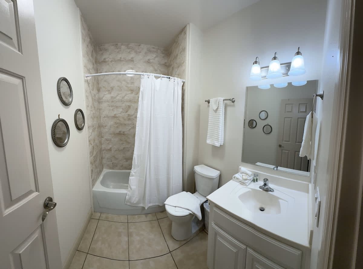 A bright bathroom with a white vanity and sink, a large mirror, and a three-light fixture above. The shower area has light-colored tiled walls and a white shower curtain, with a toilet nearby.