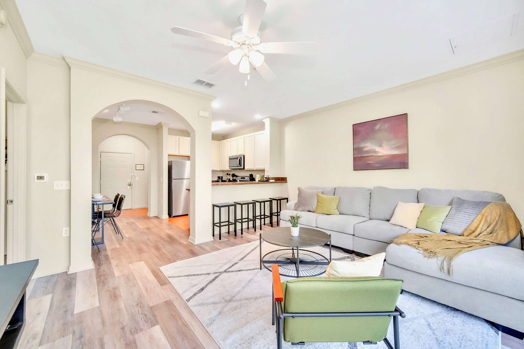 Open-concept living area flowing into the kitchen, featuring a light gray sectional sofa, a round coffee table, and a breakfast bar with stools, all on wood-look flooring.