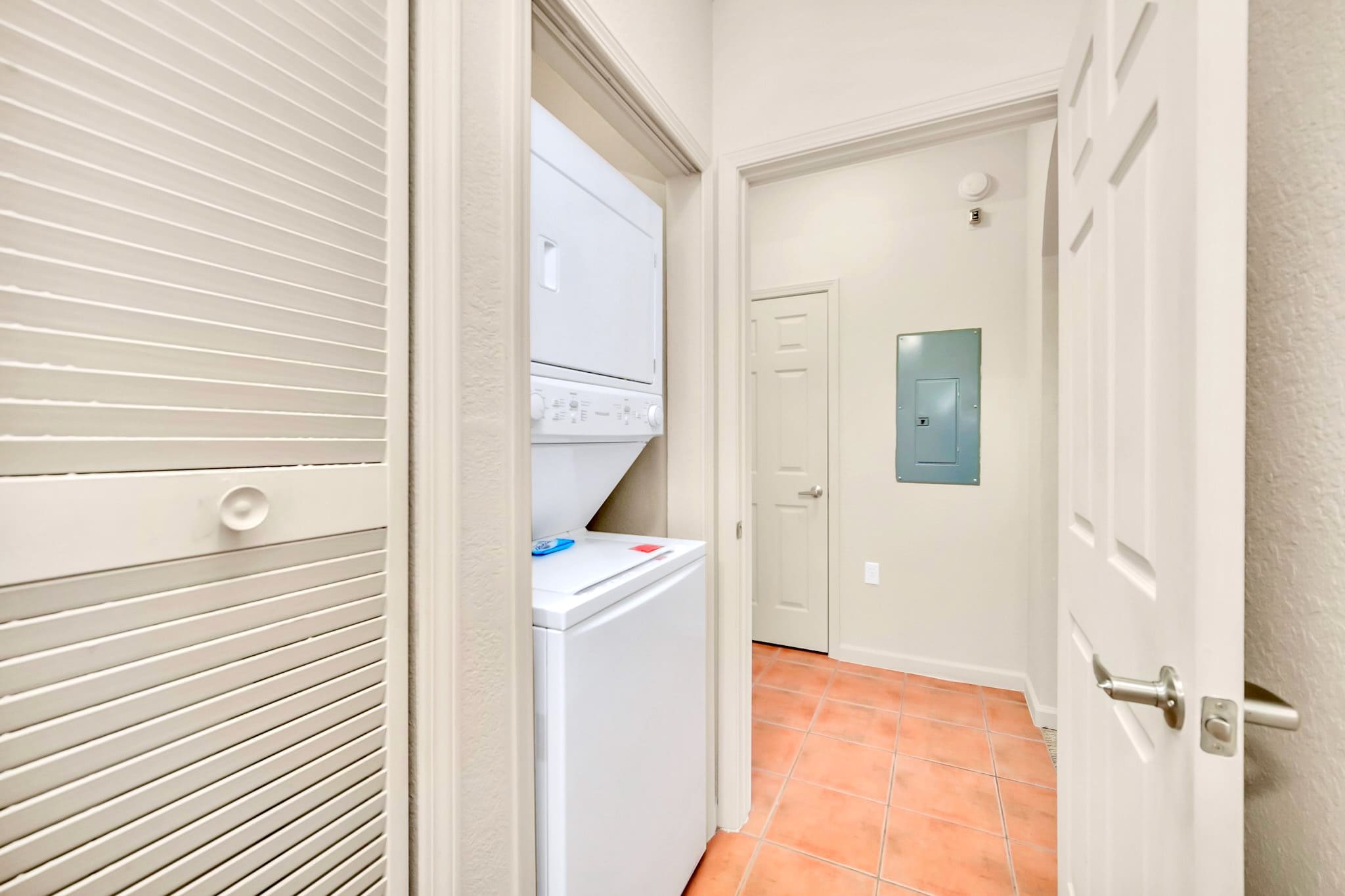 Compact laundry closet with a stacked white washer and dryer unit. The floor is tiled in orange-brown, and there's a closed louvered door to the left.