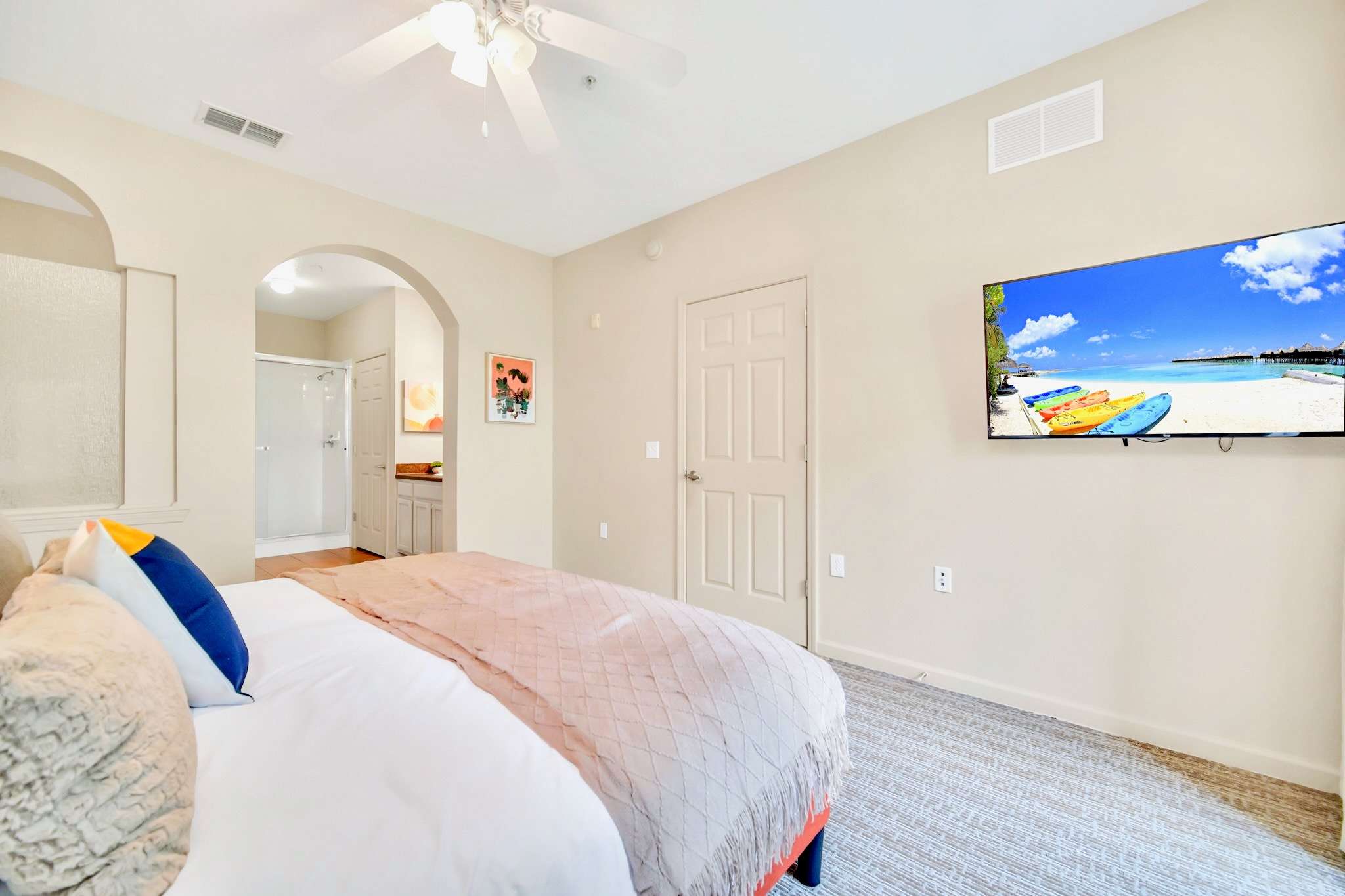 Cozy bedroom featuring a queen-sized bed with a white and peach comforter, a ceiling fan, and a wall-mounted TV displaying a beach scene. An arched doorway leads to an en-suite bathroom.