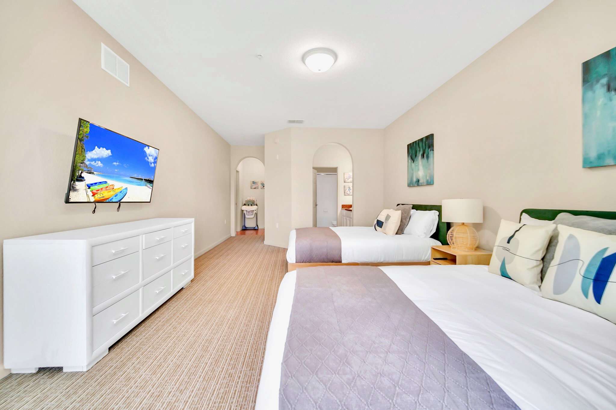 Spacious bedroom with two queen beds, a white dresser, and a wall-mounted TV. An arched opening in the background leads to a laundry area, and the room has light-colored carpet.