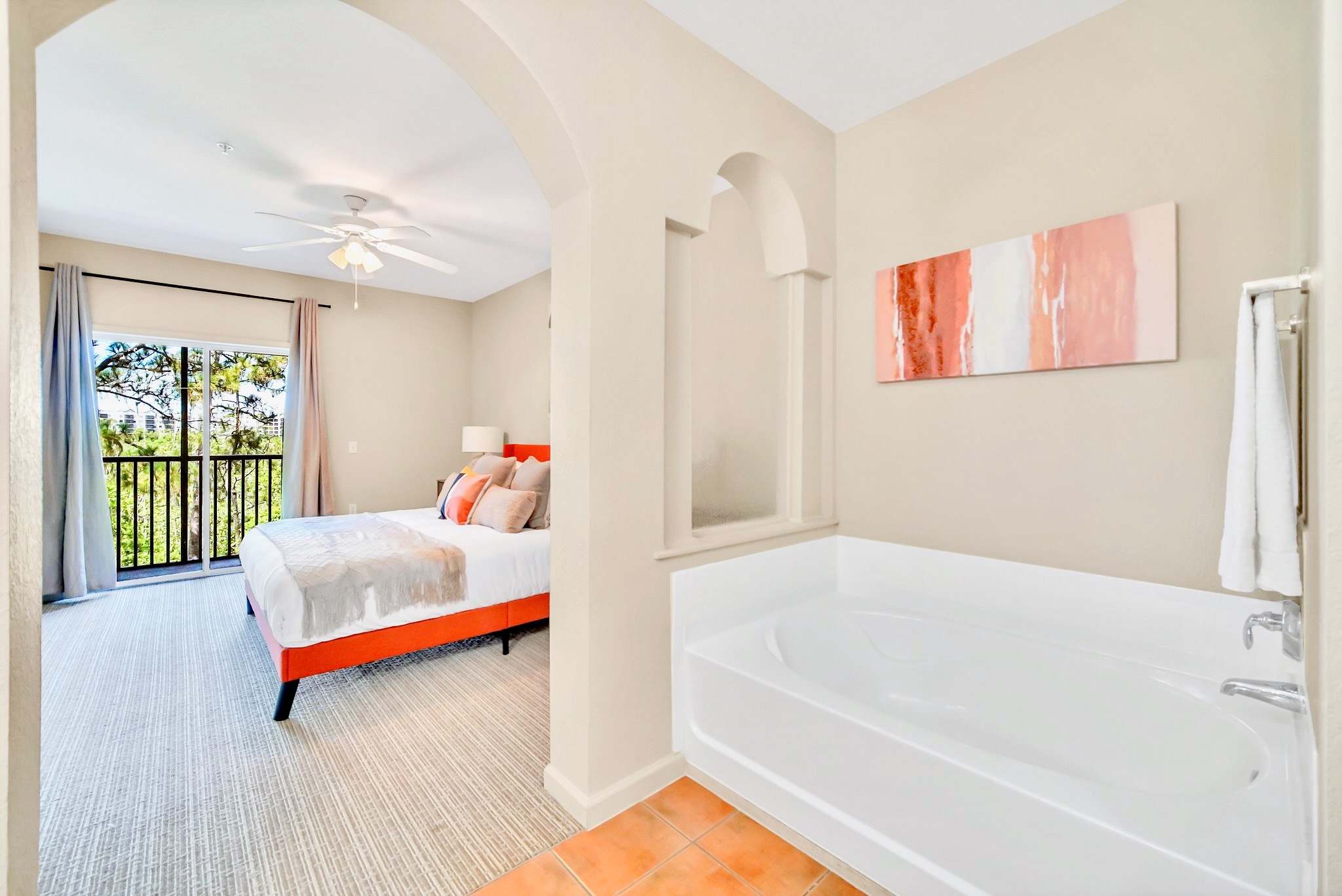 Bedroom with a queen bed, ceiling fan, and a balcony visible through glass doors. An open archway leads to a bathroom area with a large white built-in bathtub and a modern abstract painting.