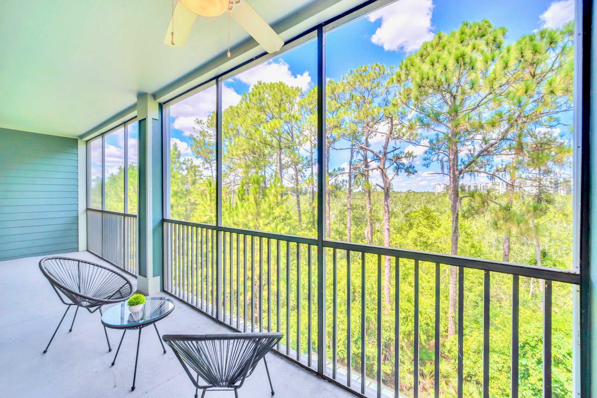Spacious screened balcony with a light green exterior wall, featuring two black wire chairs and a small glass-top table, providing panoramic views of a dense forest under a bright blue sky.