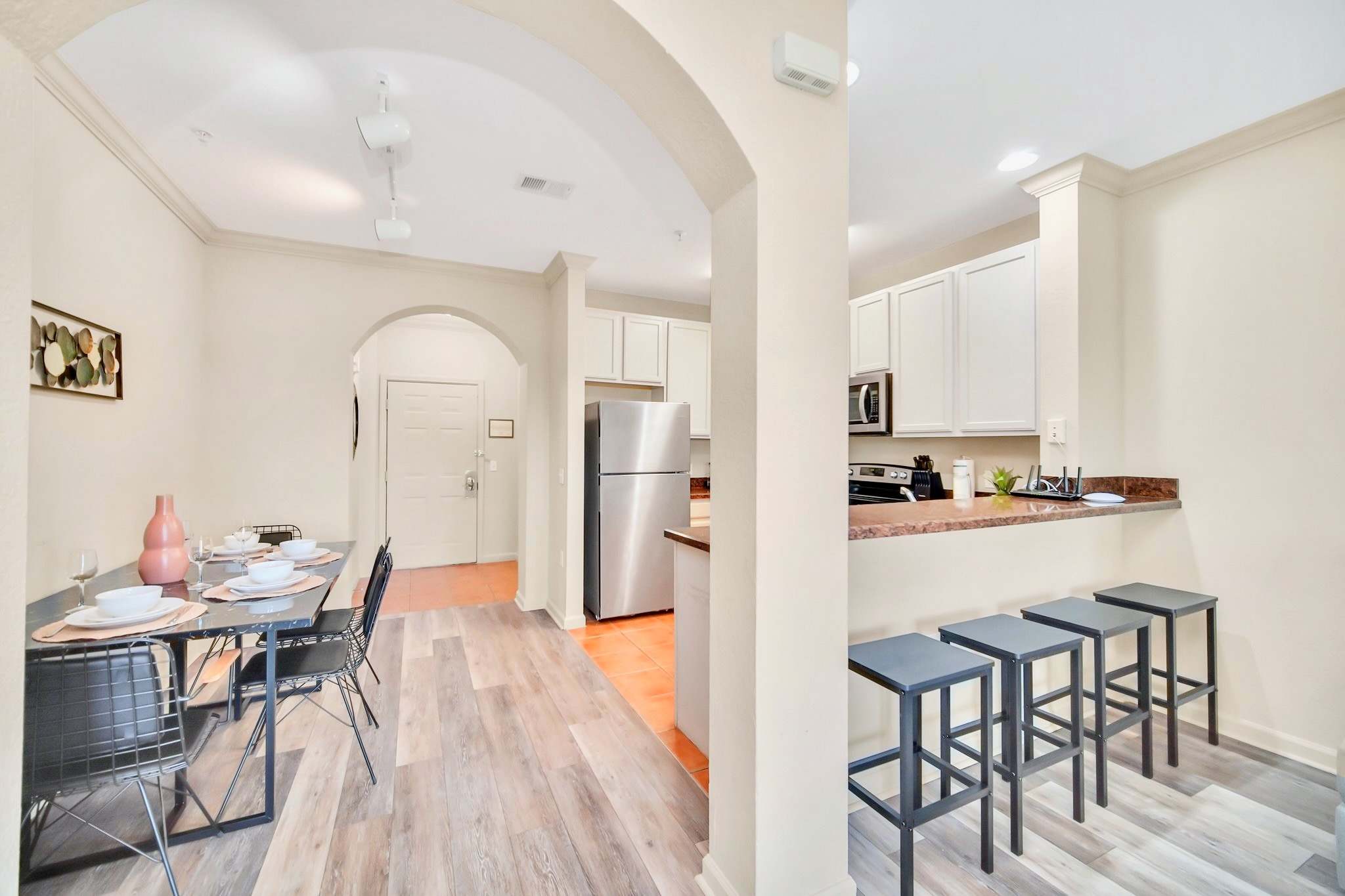 View of the dining area with a black table and chairs, adjacent to a kitchen featuring a breakfast bar with three stools, white cabinets, and stainless steel appliances.