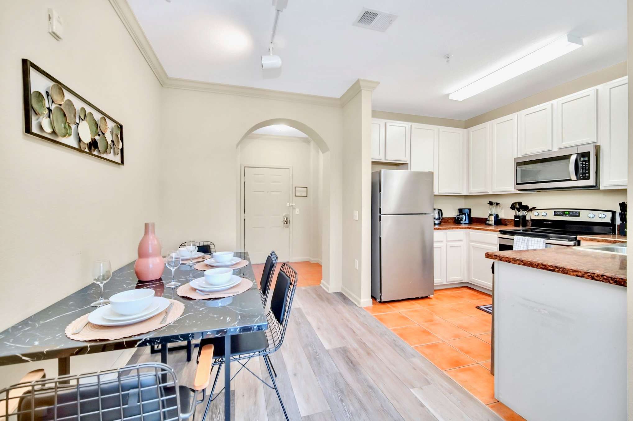 Dining area with a black marble-look table and wire chairs, leading into a kitchen with white cabinets, stainless steel appliances, and terracotta-colored tile flooring.