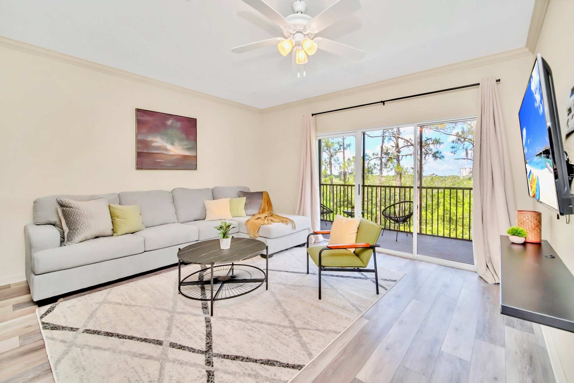 Cozy living room with a light gray sectional sofa, a round black coffee table, and a green accent chair, featuring a large sliding glass door opening to a balcony with lush green views.