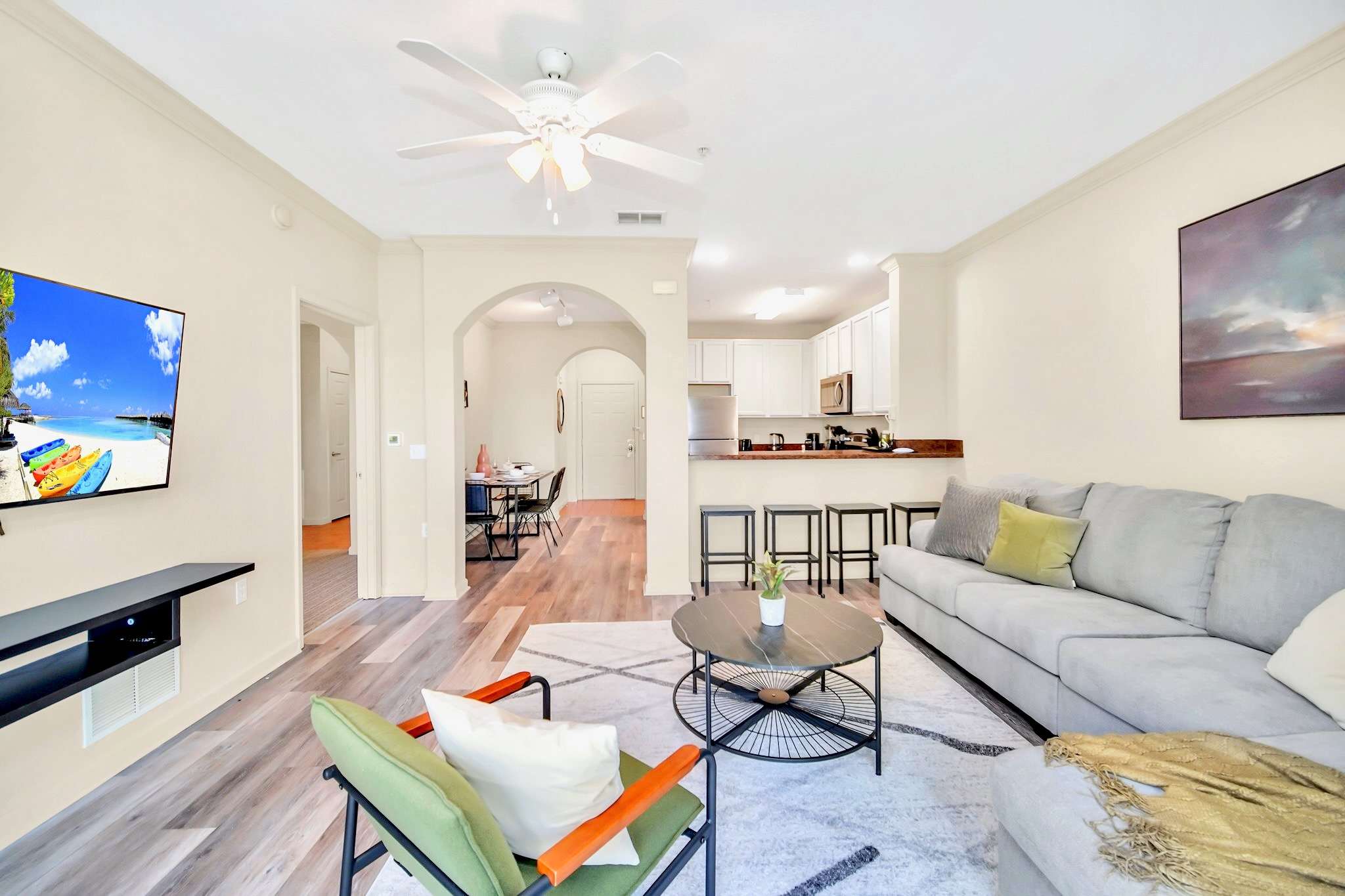 Bright living room with a light gray sectional sofa and a wall-mounted TV, offering an open view into the kitchen with a breakfast bar and the dining area through an arched doorway.