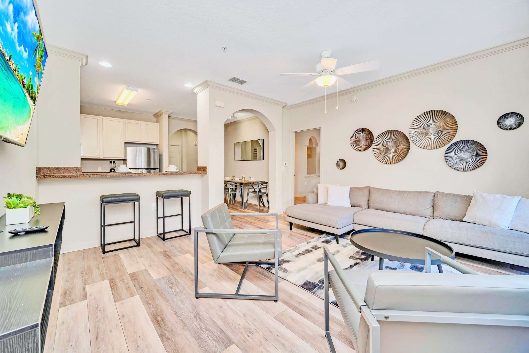 An open-concept living area with a grey L-shaped sofa, two armchairs, and a round coffee table. It connects to a kitchen bar with two stools and offers a view into the kitchen and dining area.