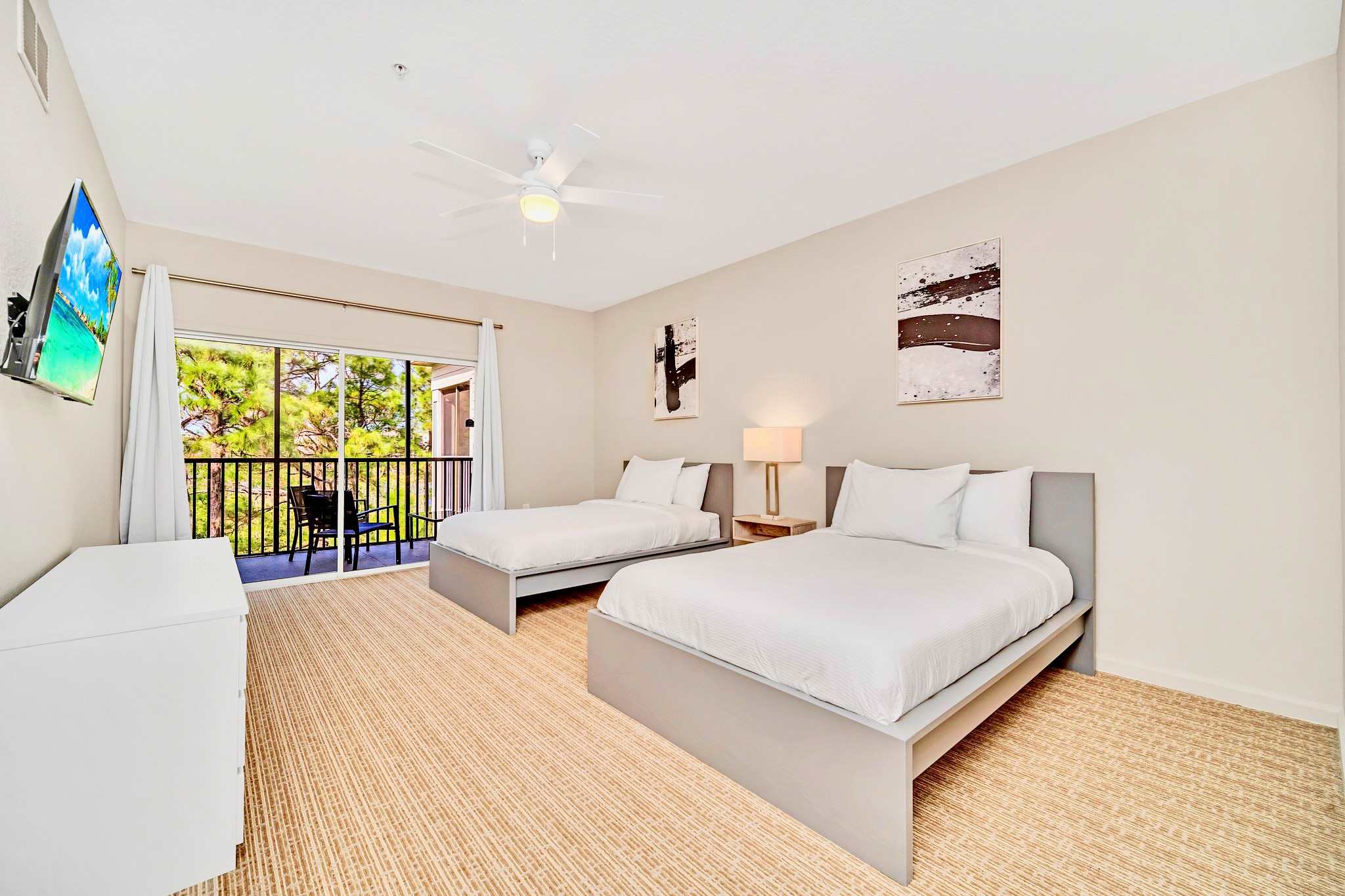 A bright bedroom featuring two single beds with grey frames and white bedding, a ceiling fan, and abstract wall art. A sliding glass door leads to a screened balcony with views of trees.