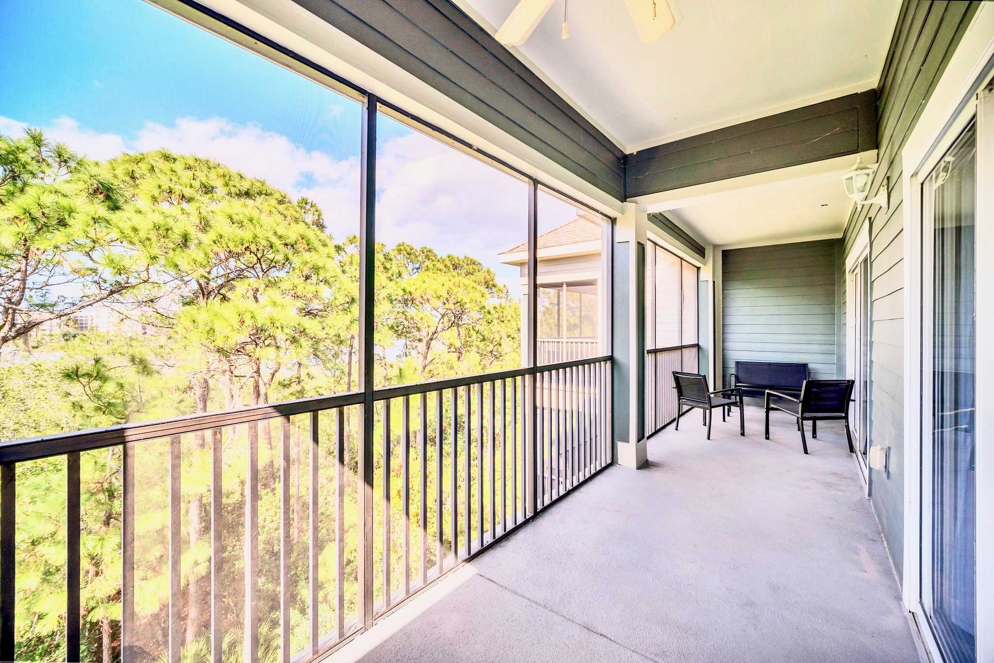 A wide screened-in balcony with dark grey railings, offering expansive views of pine trees under a blue sky. It features outdoor seating with two chairs and a small table, and a ceiling fan.