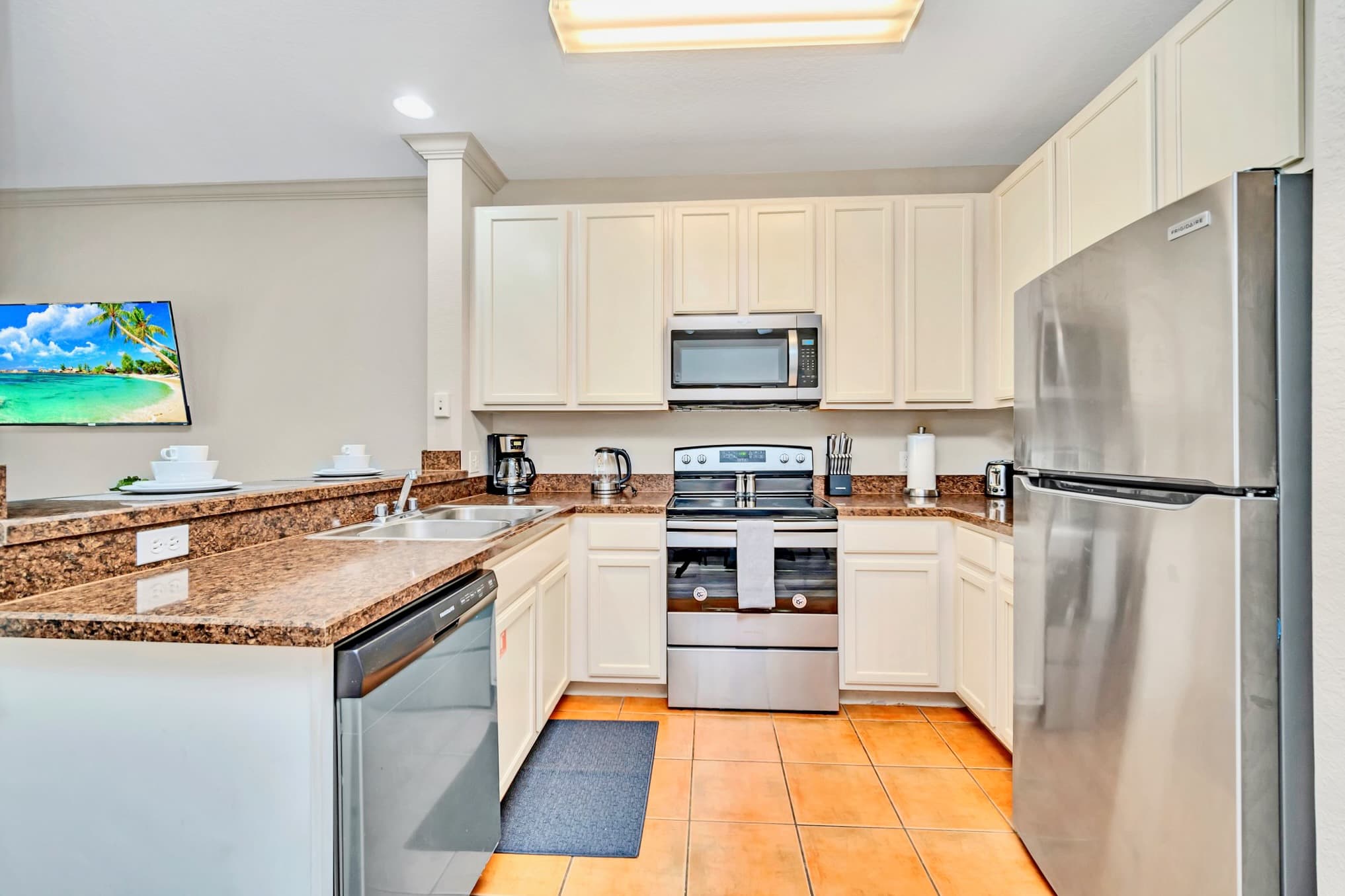 A well-equipped kitchen featuring stainless steel appliances including a refrigerator, stove, microwave, and dishwasher. It has light-colored cabinets, granite countertops, and a double sink, with terracotta-colored tile flooring.