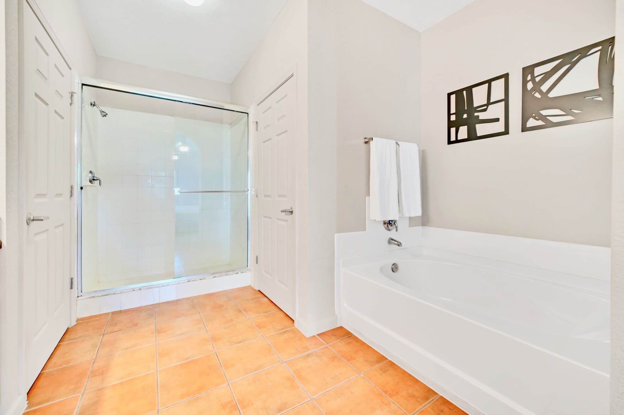 A spacious bathroom with terracotta-colored floor tiles, featuring a white built-in bathtub and a glass-enclosed walk-in shower. White towels are neatly folded on a rack above the tub.
