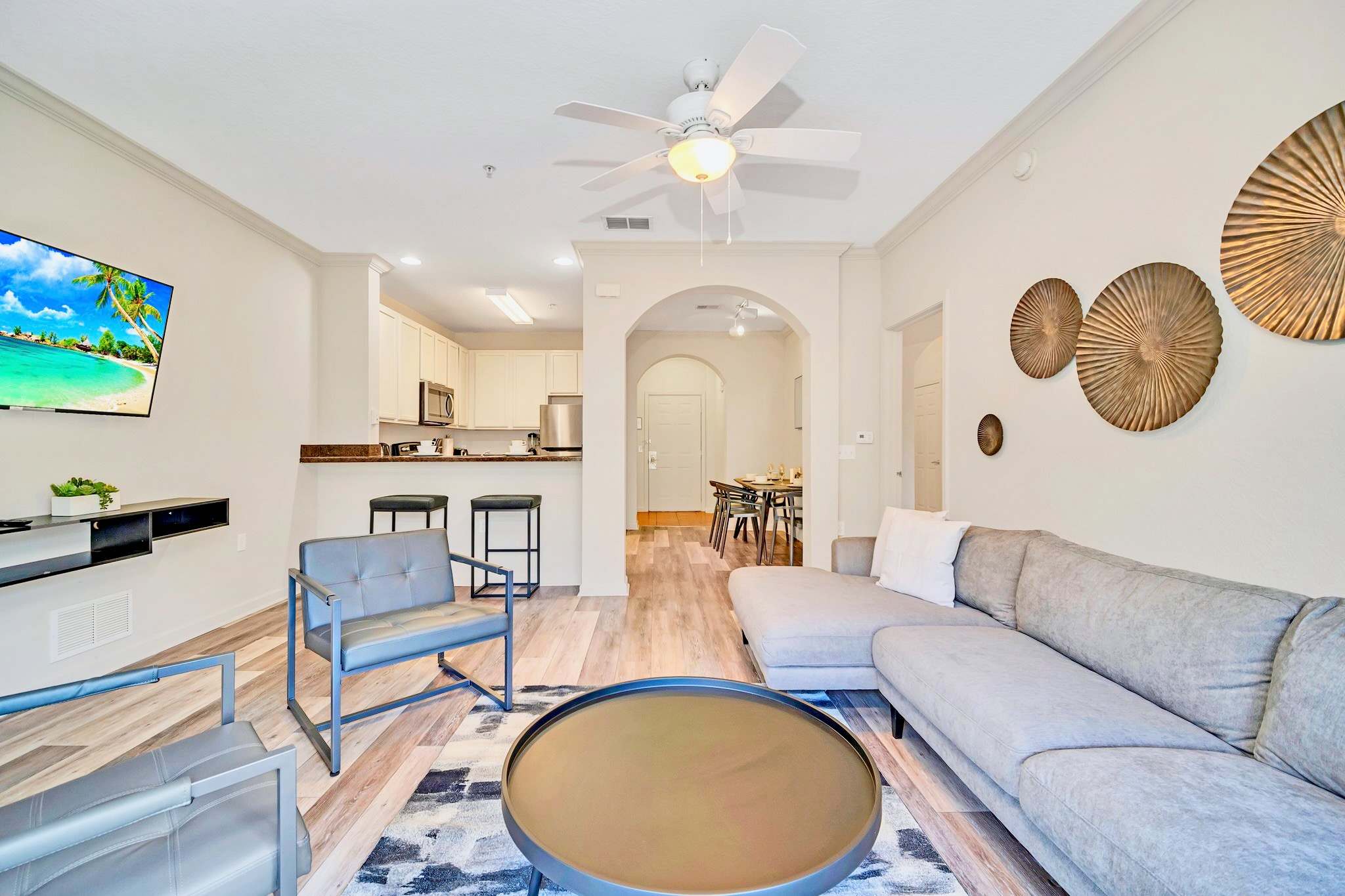 An overview of the open-concept living room, featuring a large L-shaped sofa, two grey armchairs, and a round coffee table on a patterned rug. The space flows into the kitchen area with a breakfast bar and the dining room beyond.