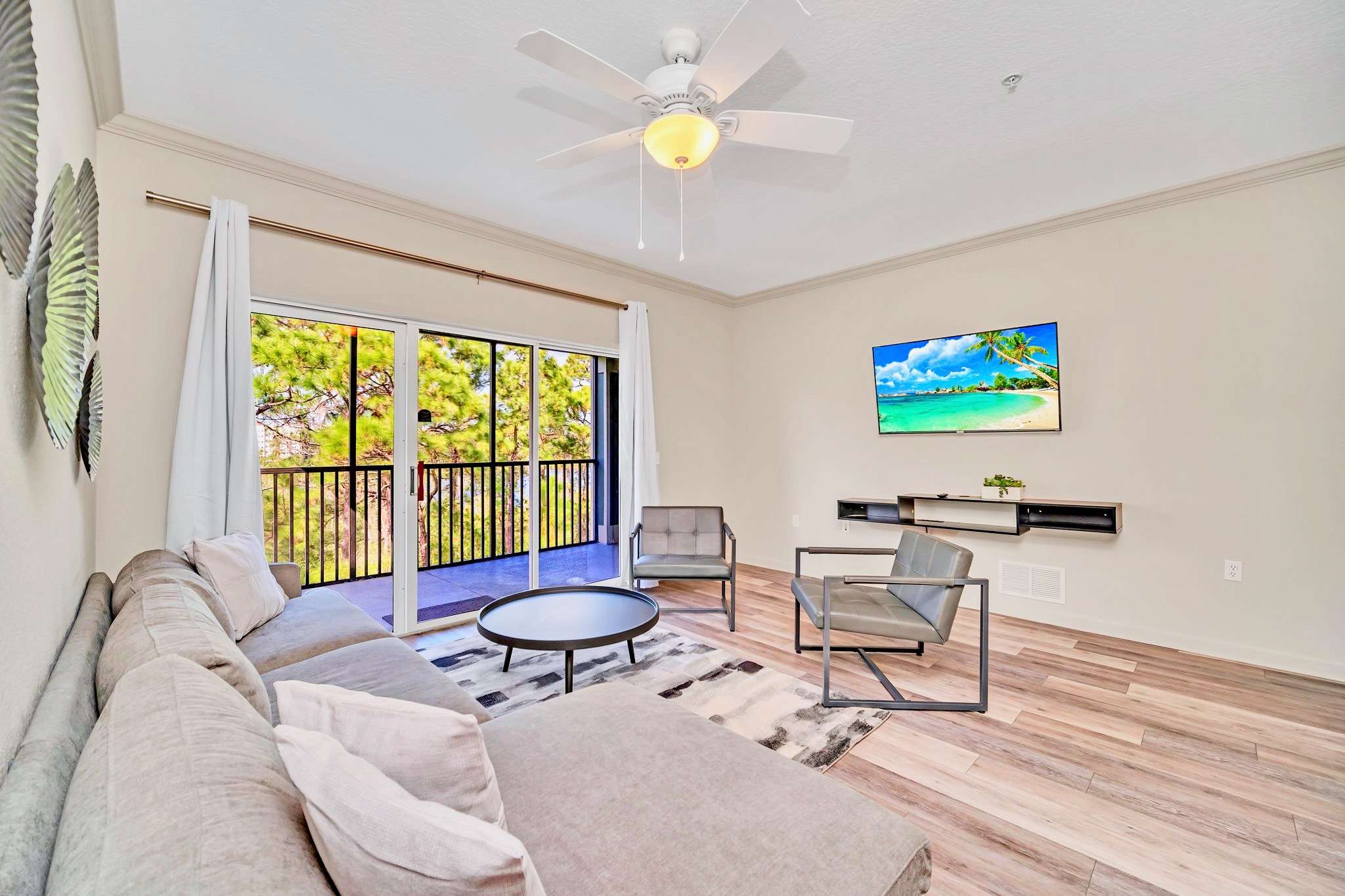 A bright living room with a large L-shaped sectional sofa, two grey armchairs, and a round coffee table on a patterned rug. A wall-mounted TV shows a tropical scene, and sliding glass doors lead to a balcony with tree views.