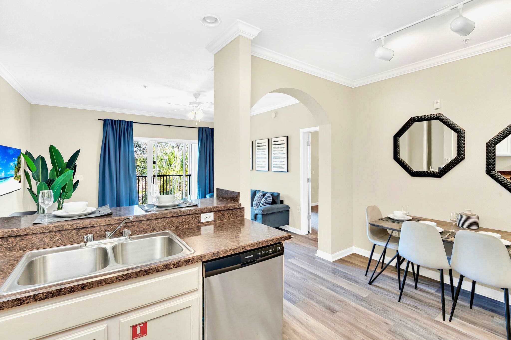 Kitchen view showing a double sink and dishwasher integrated into the dark countertop, with light cabinets below. The open layout provides a glimpse into the living room and dining area.