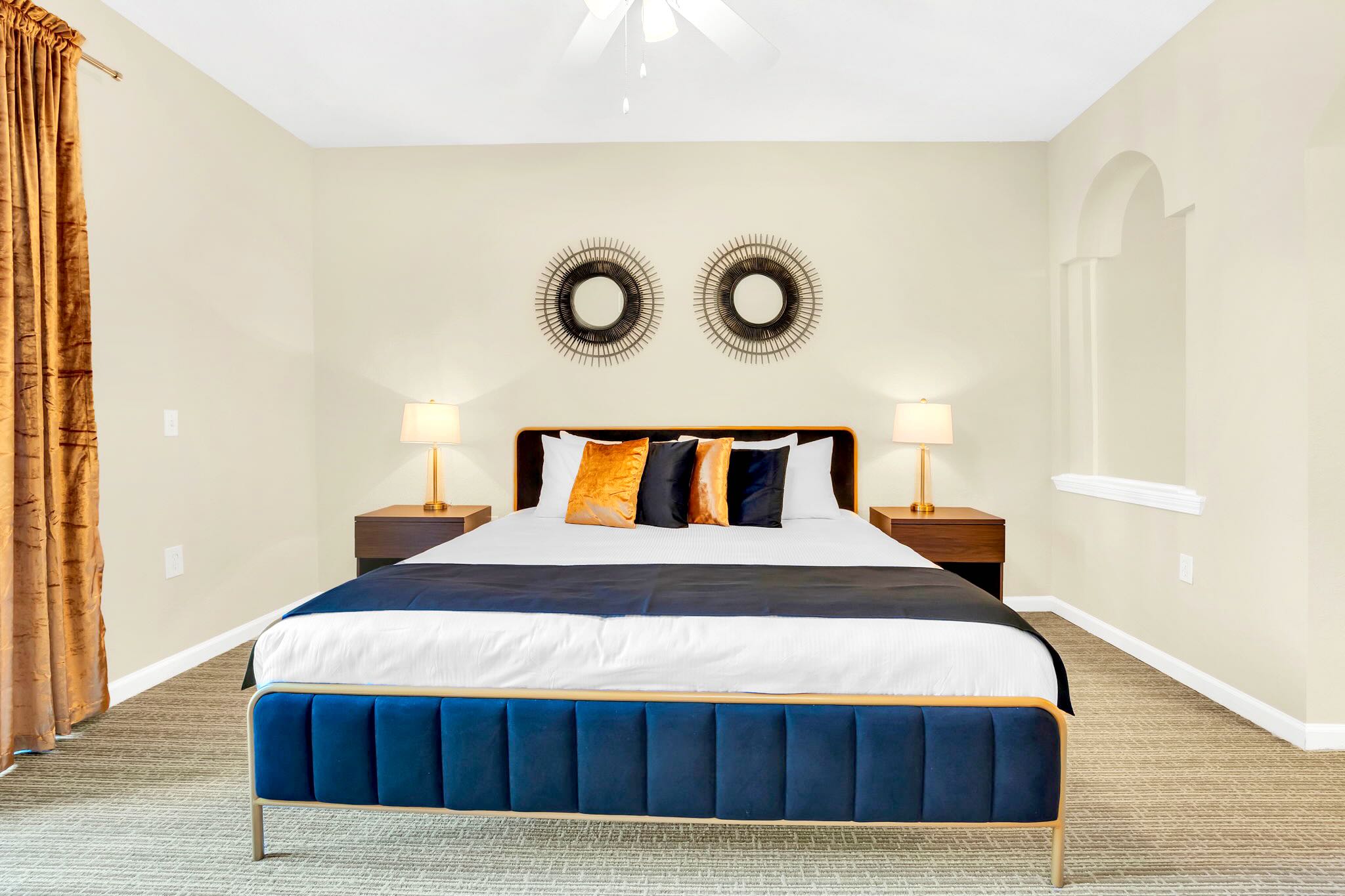 This bright bedroom features a king-sized bed with a blue upholstered frame, two wooden nightstands with gold lamps, and two circular sunburst wall decorations above the headboard. A ceiling fan is visible overhead.