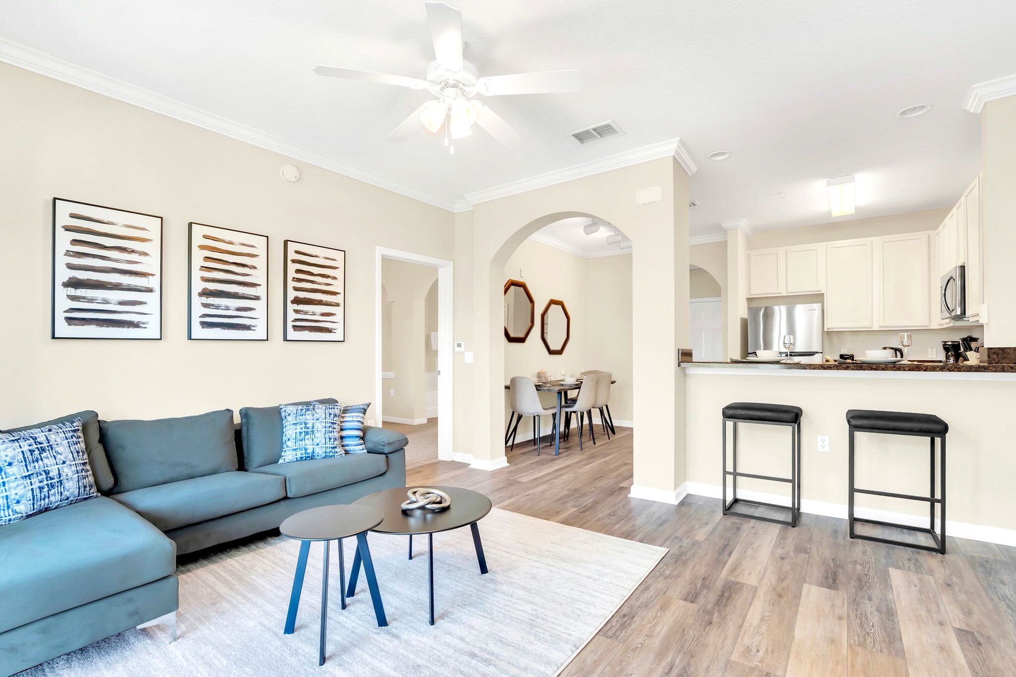 Open-concept living space featuring a blue sectional sofa, two round coffee tables, and an area rug, with a view into the dining area and kitchen through arched doorways. A ceiling fan is visible overhead.