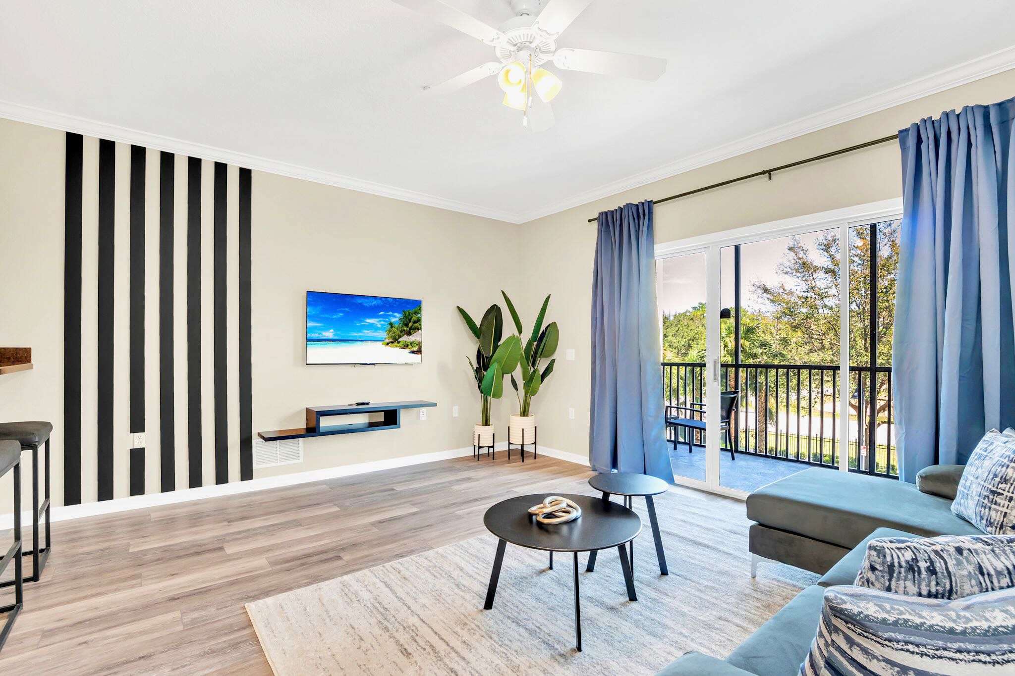 Bright living room with a blue sectional sofa, two round black coffee tables, and a wall-mounted TV. Large sliding glass doors lead to a balcony, flanked by tall green potted plants.