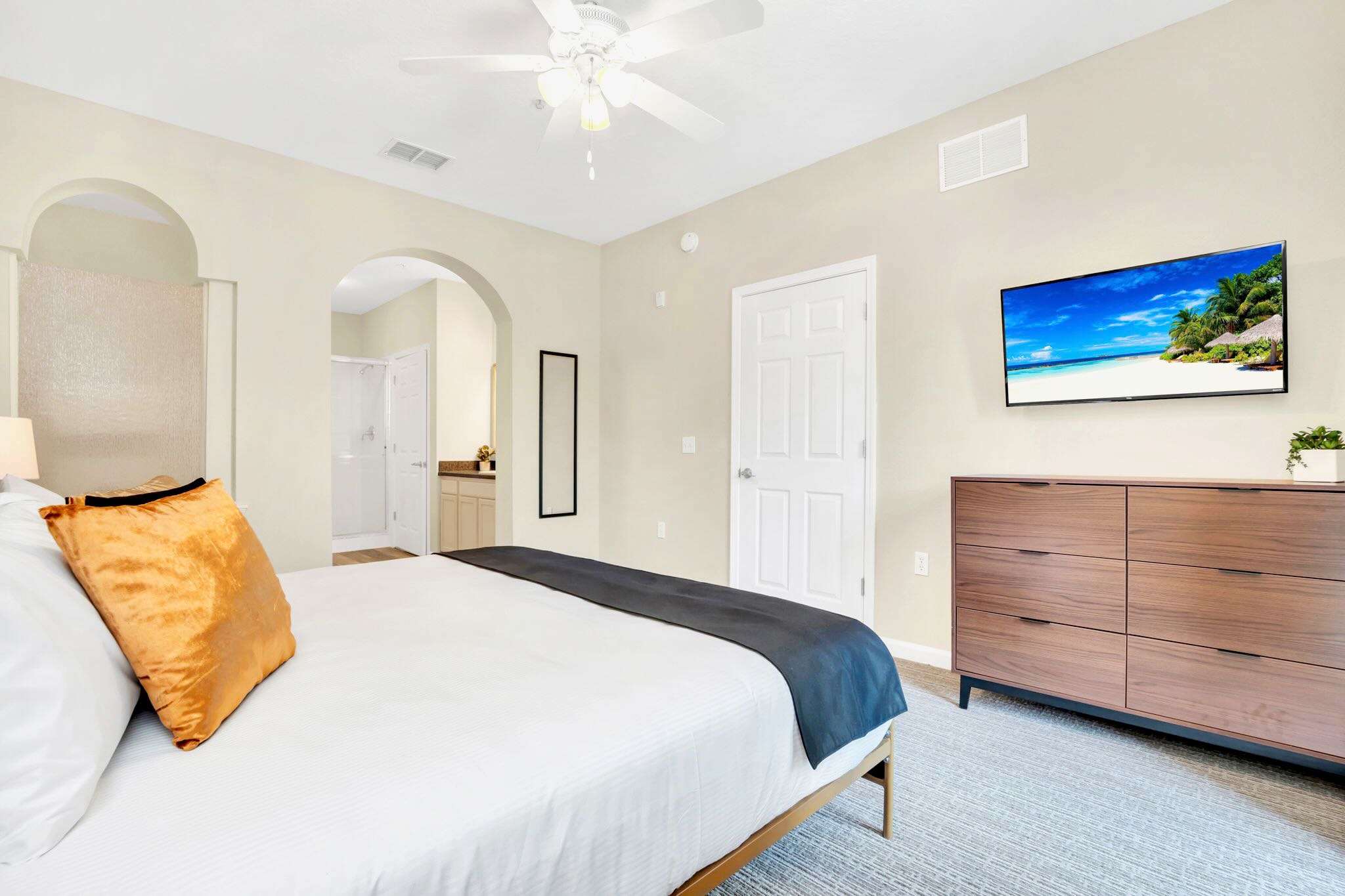 A different view of the bedroom shows the king-sized bed with a dark blue runner, a dark wood dresser with a wall-mounted TV, and an arched doorway leading to an en-suite bathroom.
