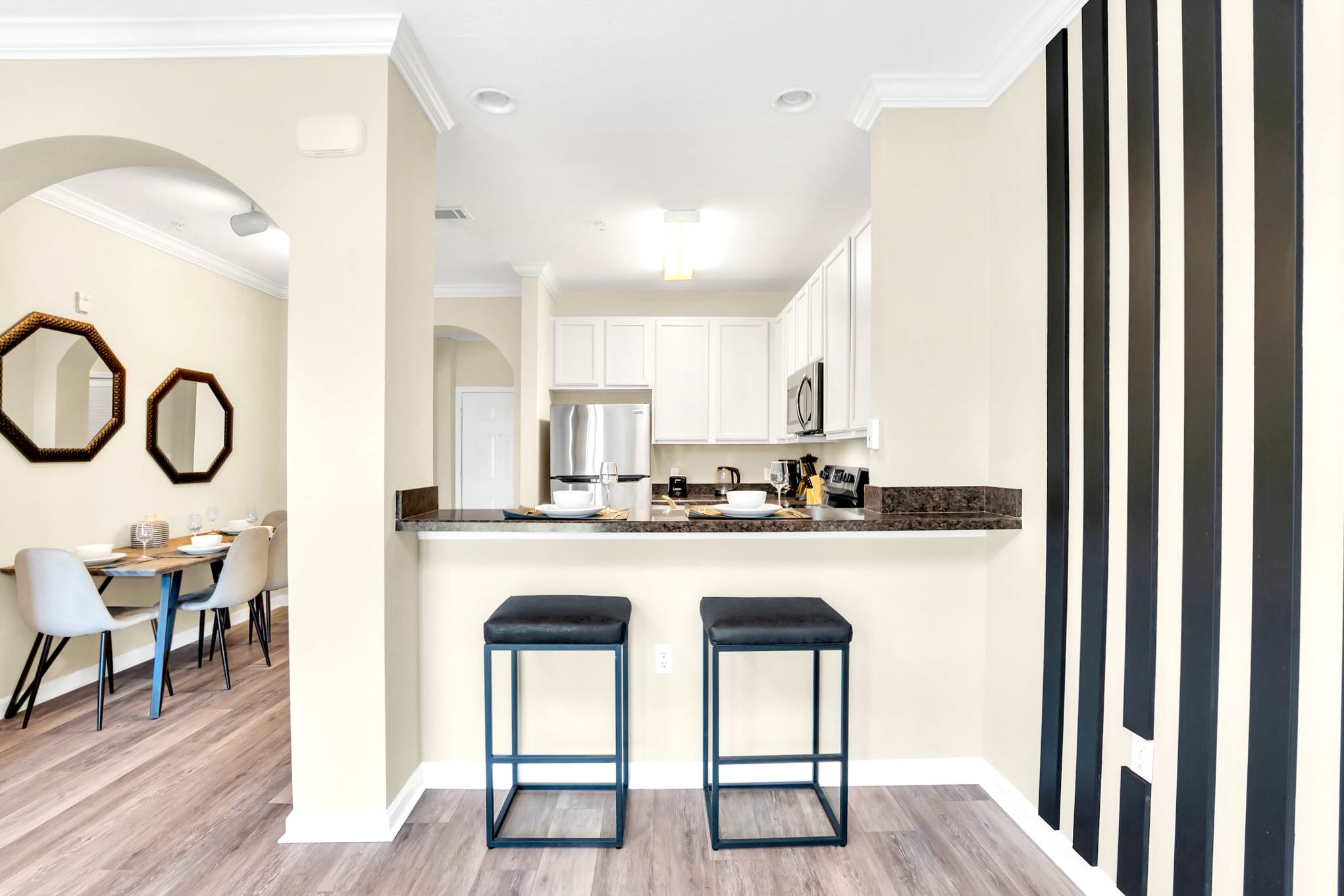 View of the kitchen's breakfast bar from the living area, featuring two black bar stools and a dark countertop. The kitchen with light cabinets is visible beyond, and the dining area is to the left through an archway.