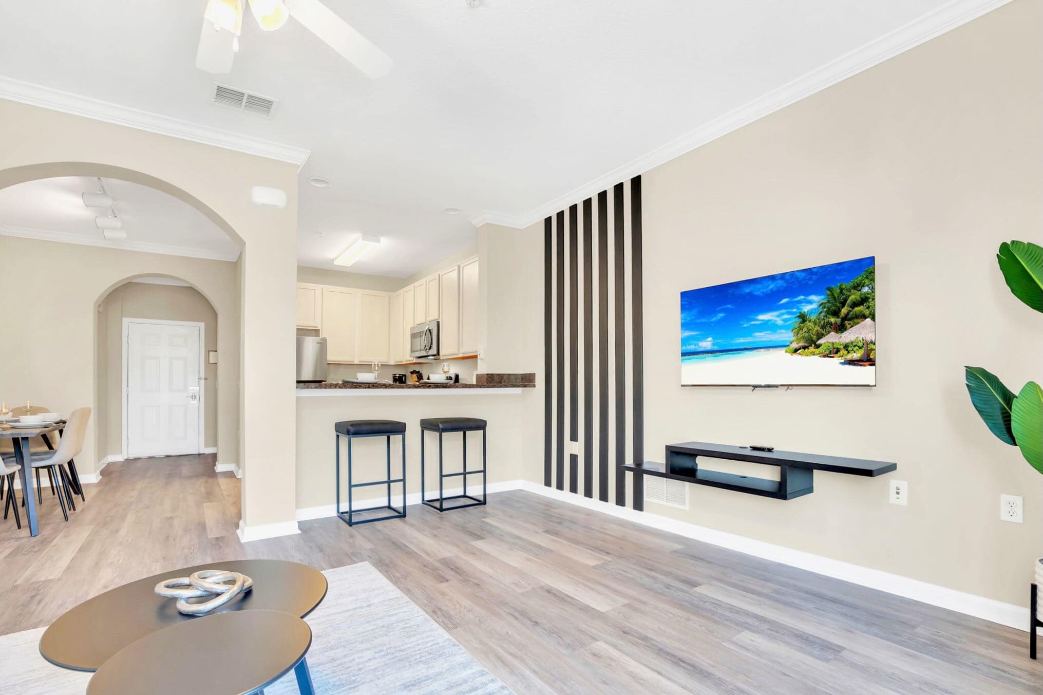Living room with a wall-mounted flat-screen TV above a floating shelf, set against a feature wall with vertical black stripes. A breakfast bar with two black stools connects to the kitchen.
