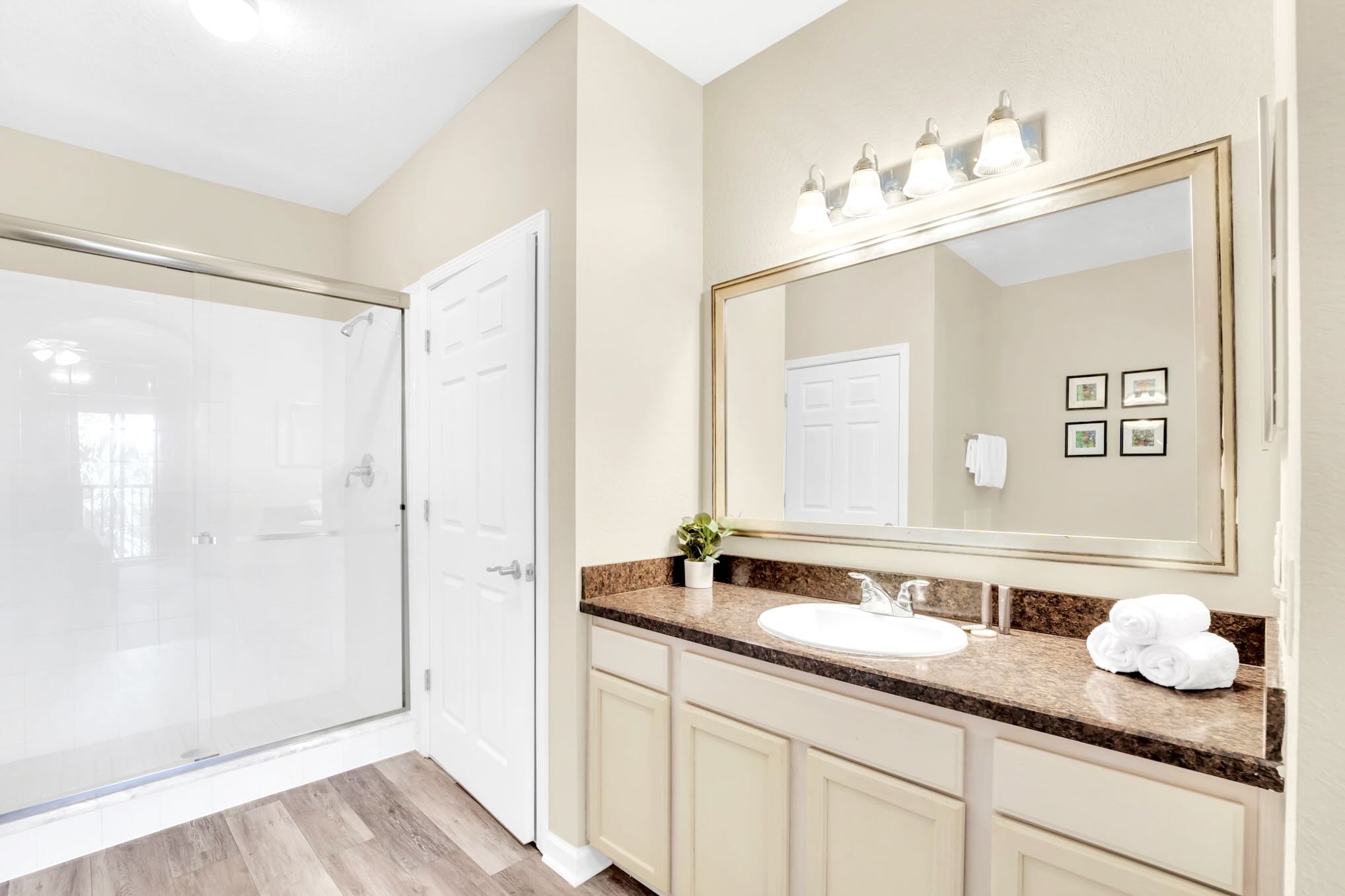 The bathroom vanity features a single white sink set into a brown granite-look countertop with light-colored cabinets below. A large framed mirror hangs above, reflecting a walk-in shower with a glass door.
