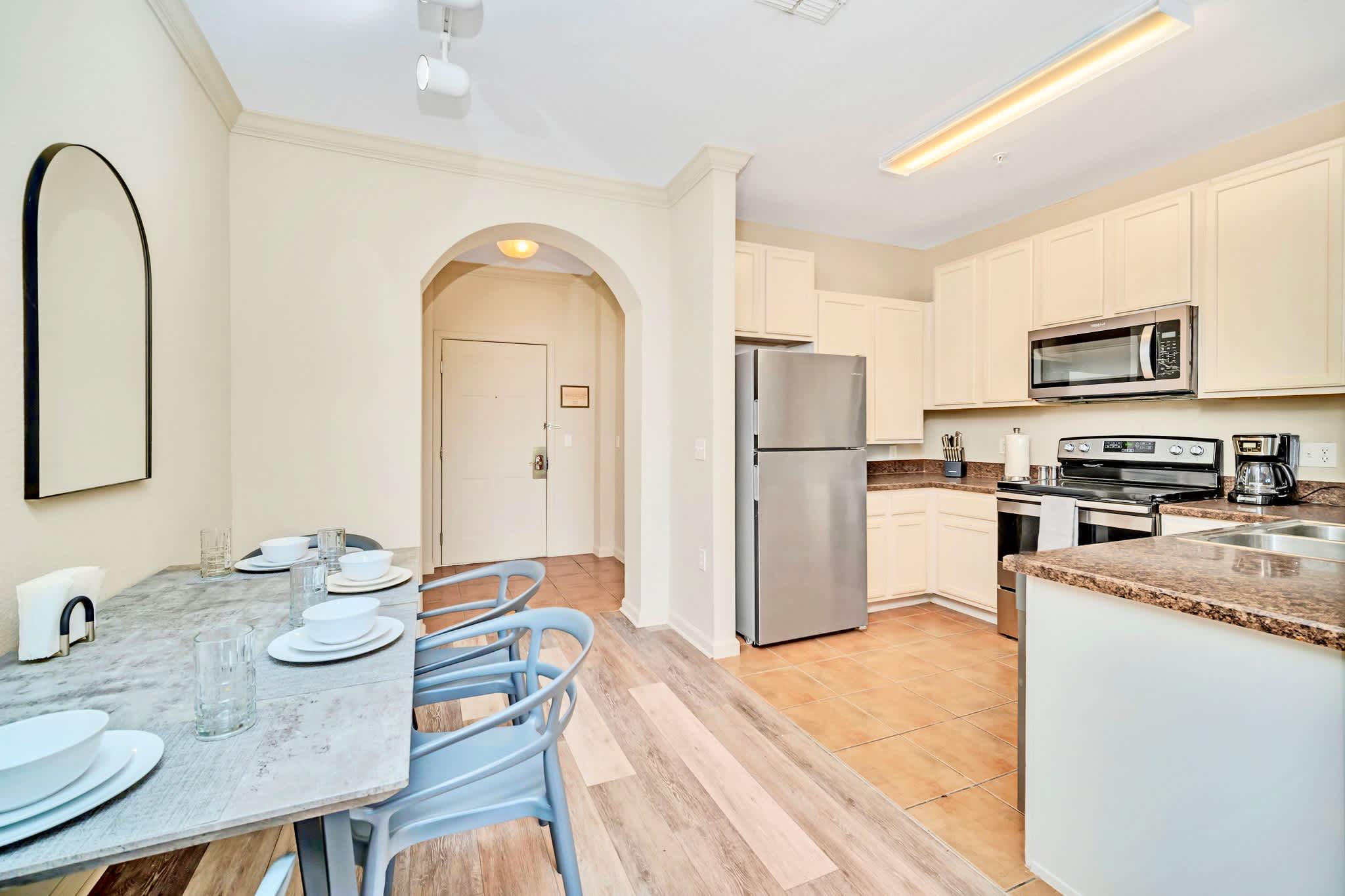 Dining area with a gray rectangular table and chairs, leading into a kitchen with stainless steel appliances and light cabinetry, with the front entrance door visible in the background.