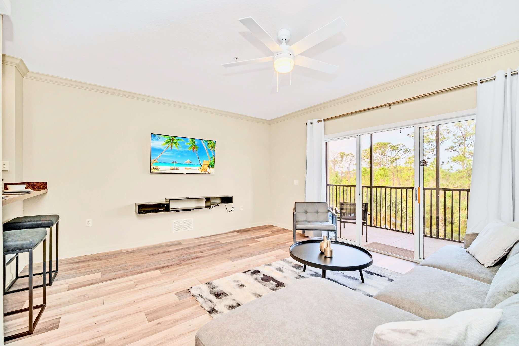 Bright living room with a light gray sectional sofa, a round coffee table, and a wall-mounted TV, adjacent to a breakfast bar with two stools, all on light wood-look flooring.