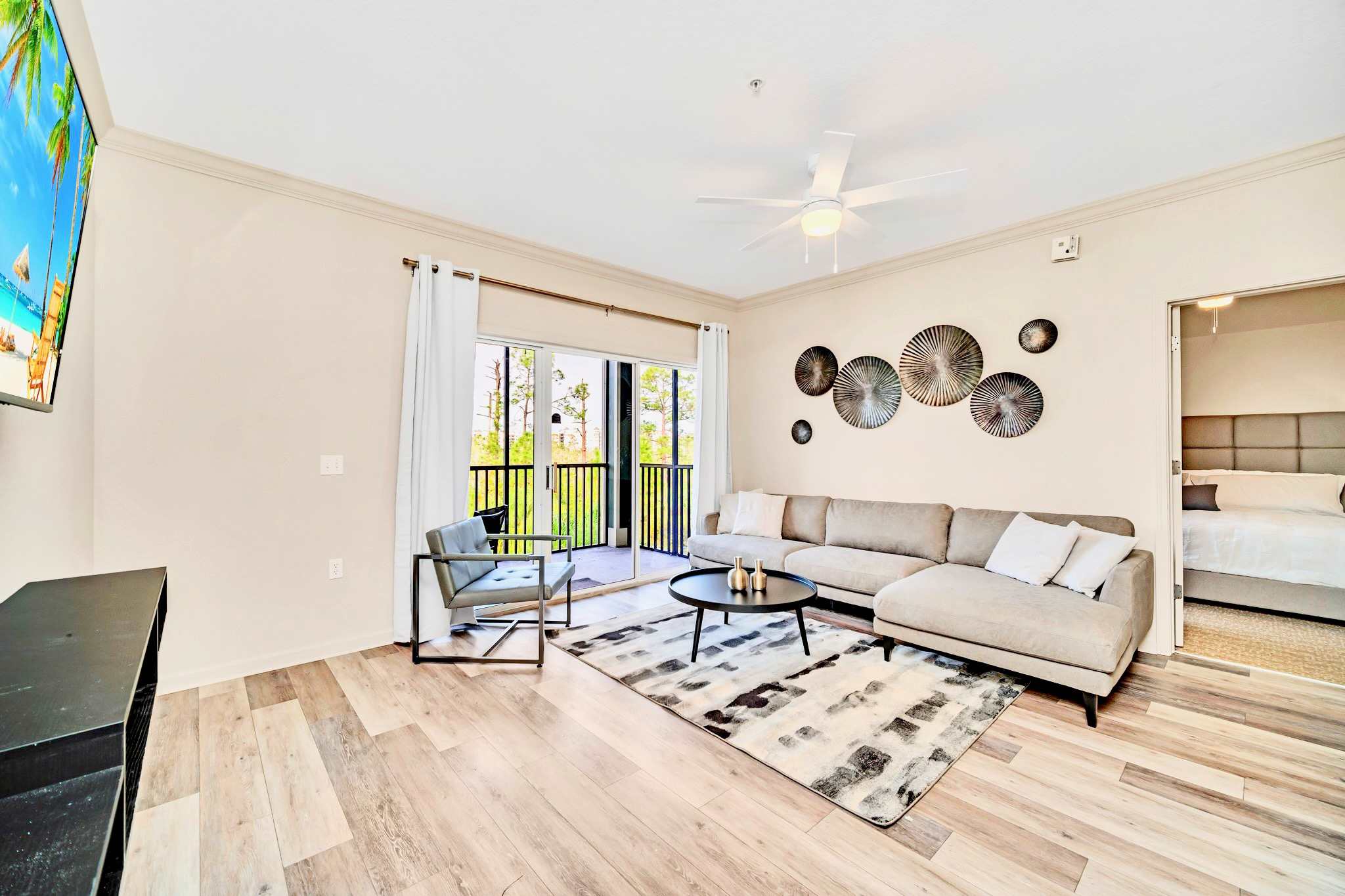 Modern living room featuring a light gray sectional sofa, a round coffee table, and a large wall-mounted TV, with light wood-look flooring and access to a balcony.