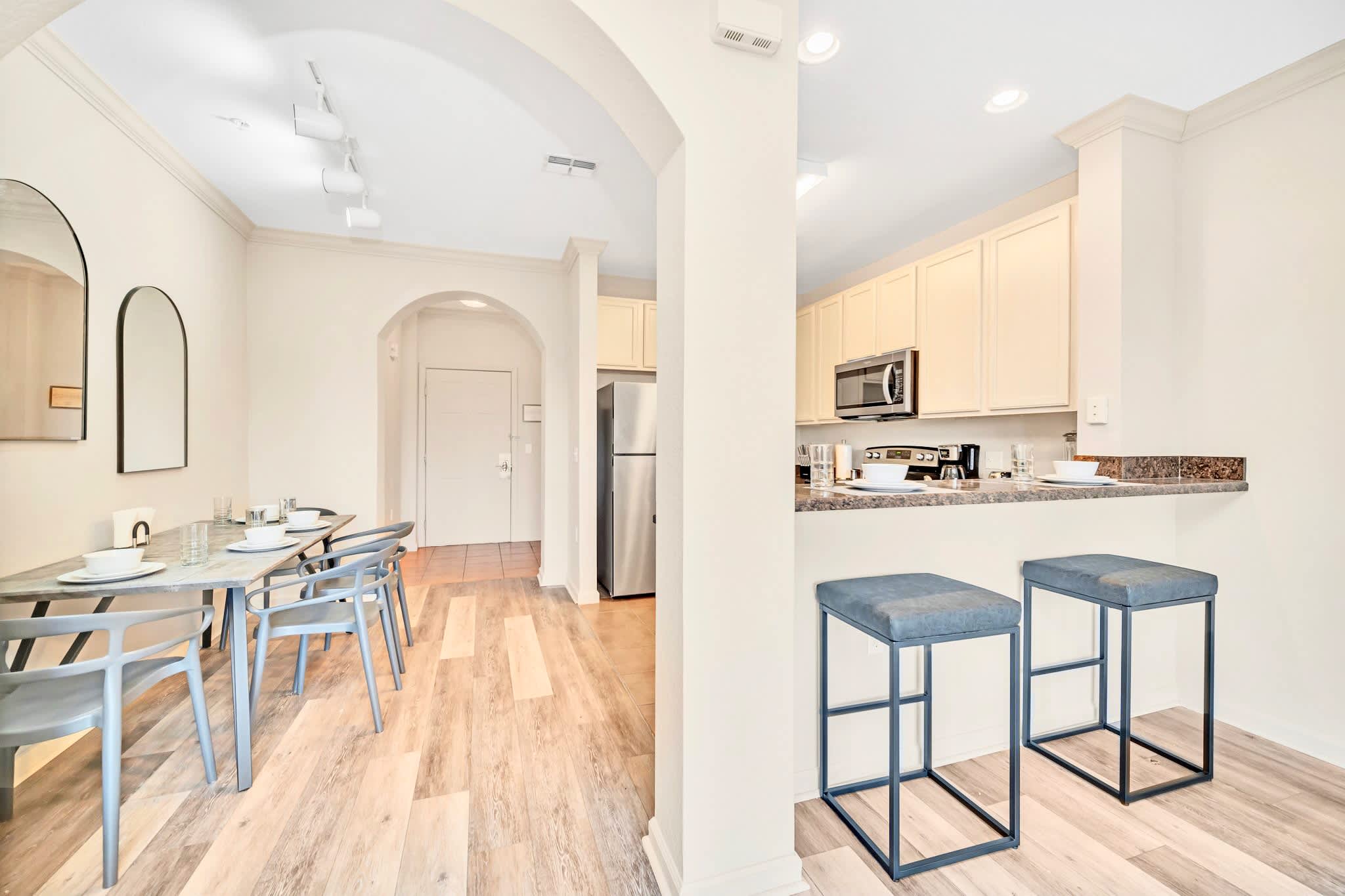 An open-concept dining area with a light wood table and blue chairs, flowing into a kitchen with cream cabinets, stainless steel appliances, and a breakfast bar with two blue stools. Light wood-look flooring connects both spaces.