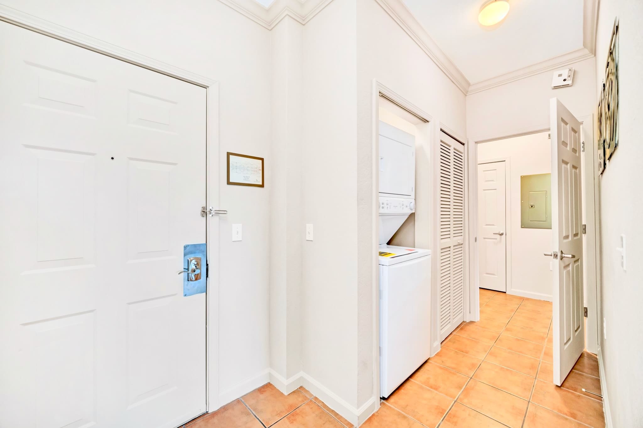 A bright entryway with terracotta-colored floor tiles, featuring a stacked washer and dryer unit in a closet, and a white front door. The space is clean and functional, providing immediate access to laundry facilities.