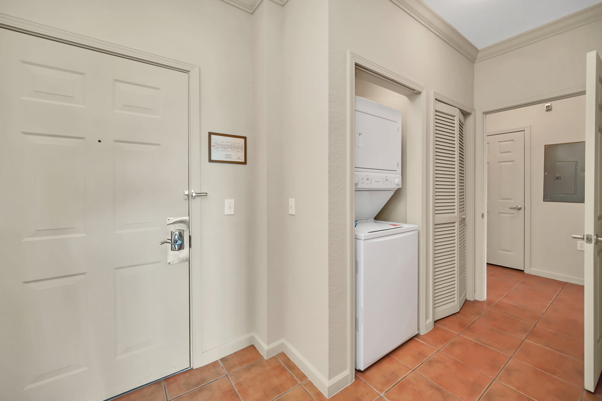 An entryway with terracotta tile flooring, a white front door, and a closet housing a stacked white washer and dryer unit.